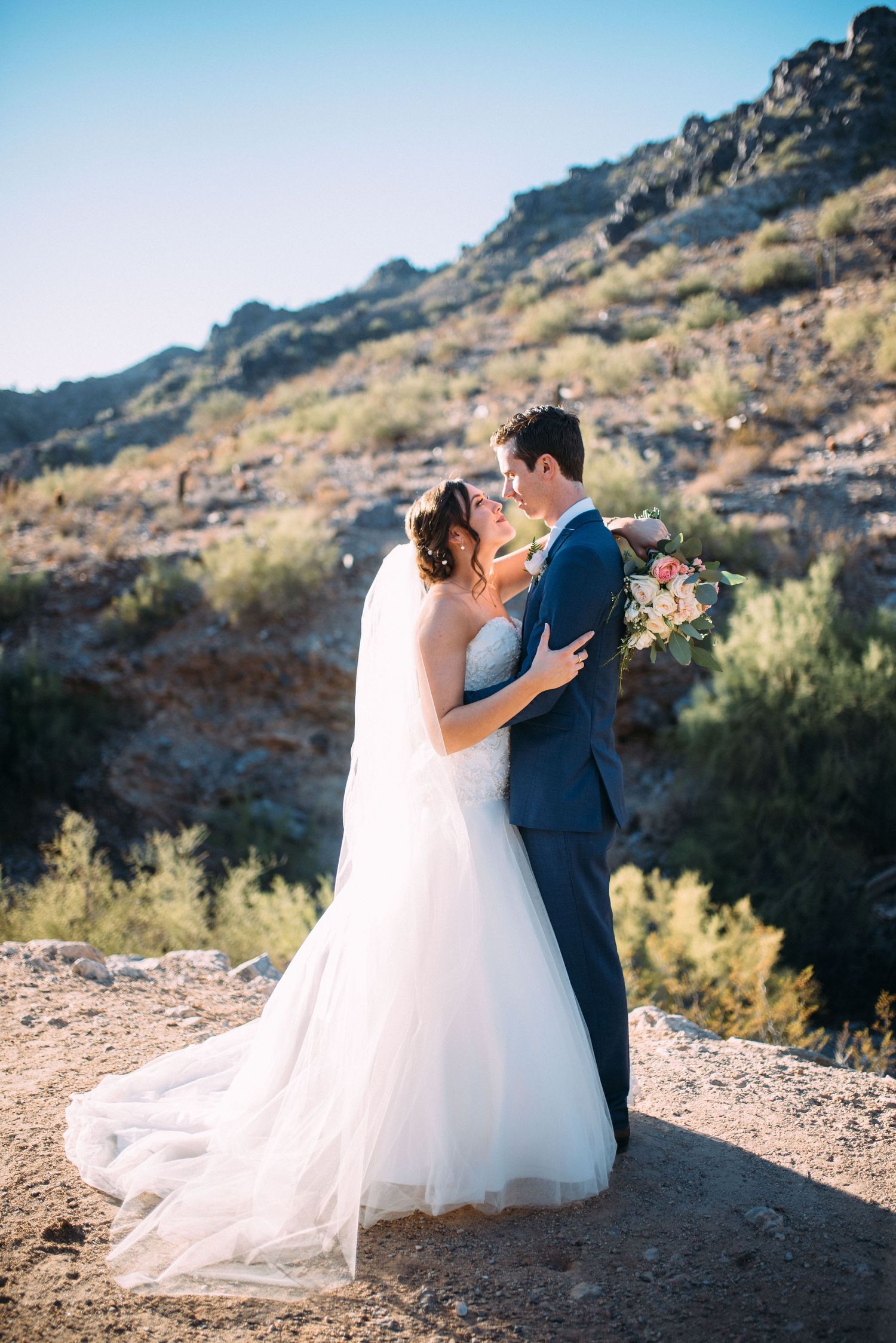 Newlyweds share a romantic kiss against a scenic desert mountain backdrop at sunset.