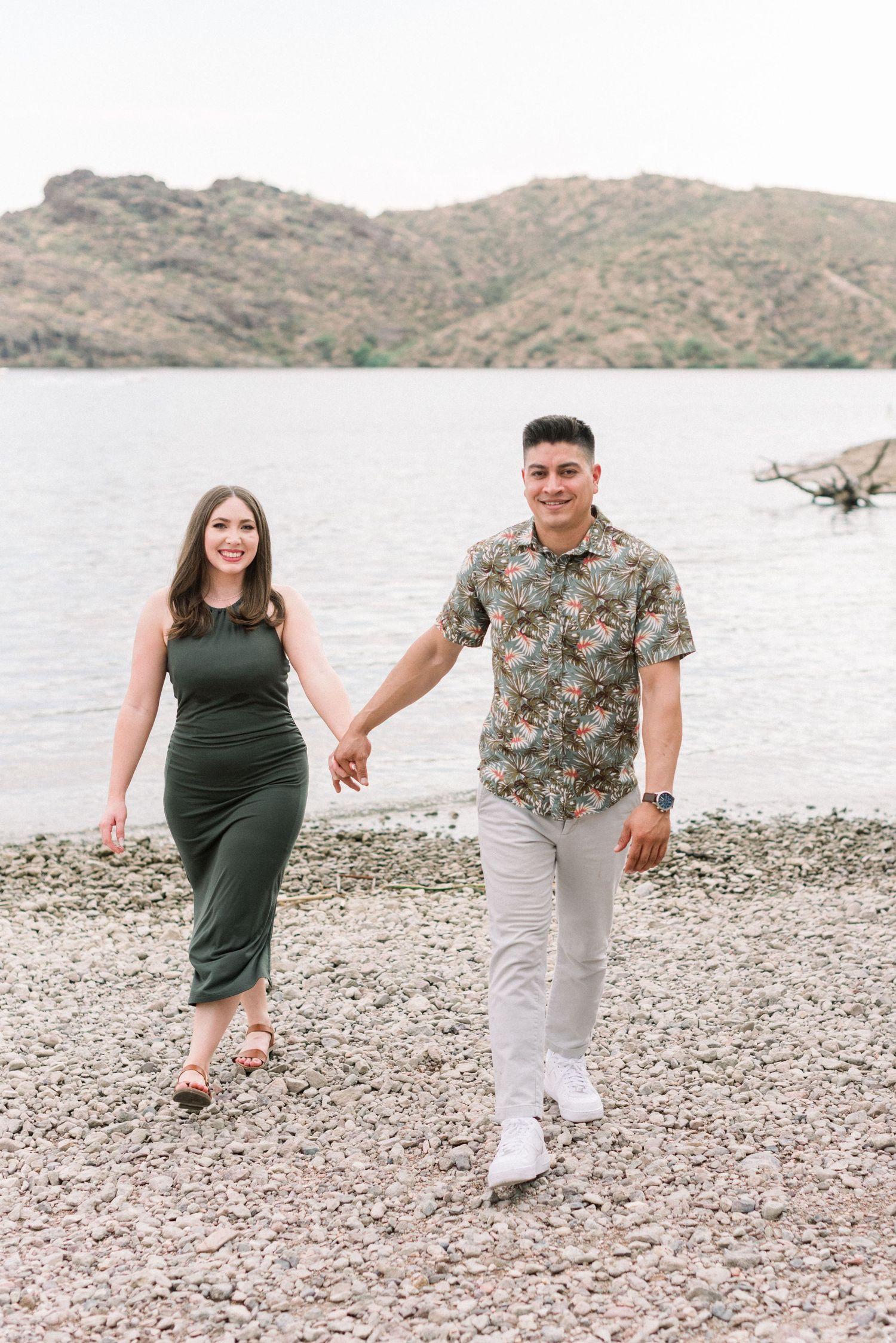 Couple walking hand in hand on rocky shoreline with mountain landscape in background.