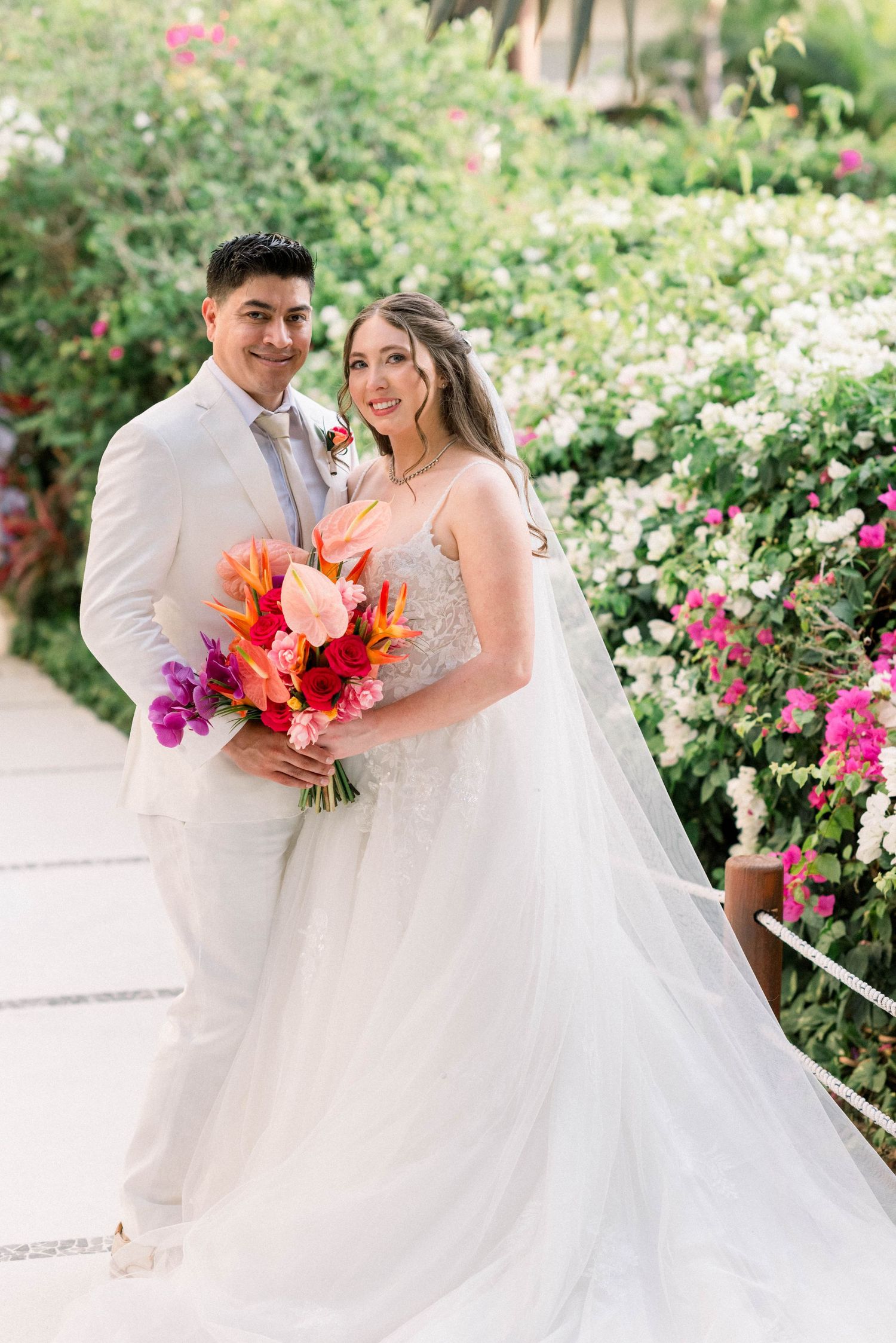 A wedding couple poses in a lush garden with vibrant coral and purple florals against white blooms.