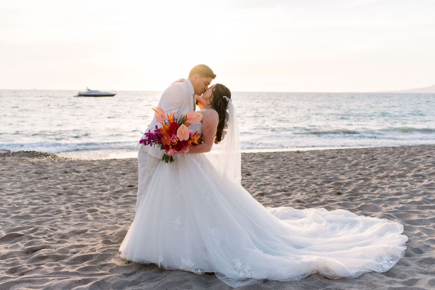 Wedding couple embracing on beach with colorful bouquet at sunset.