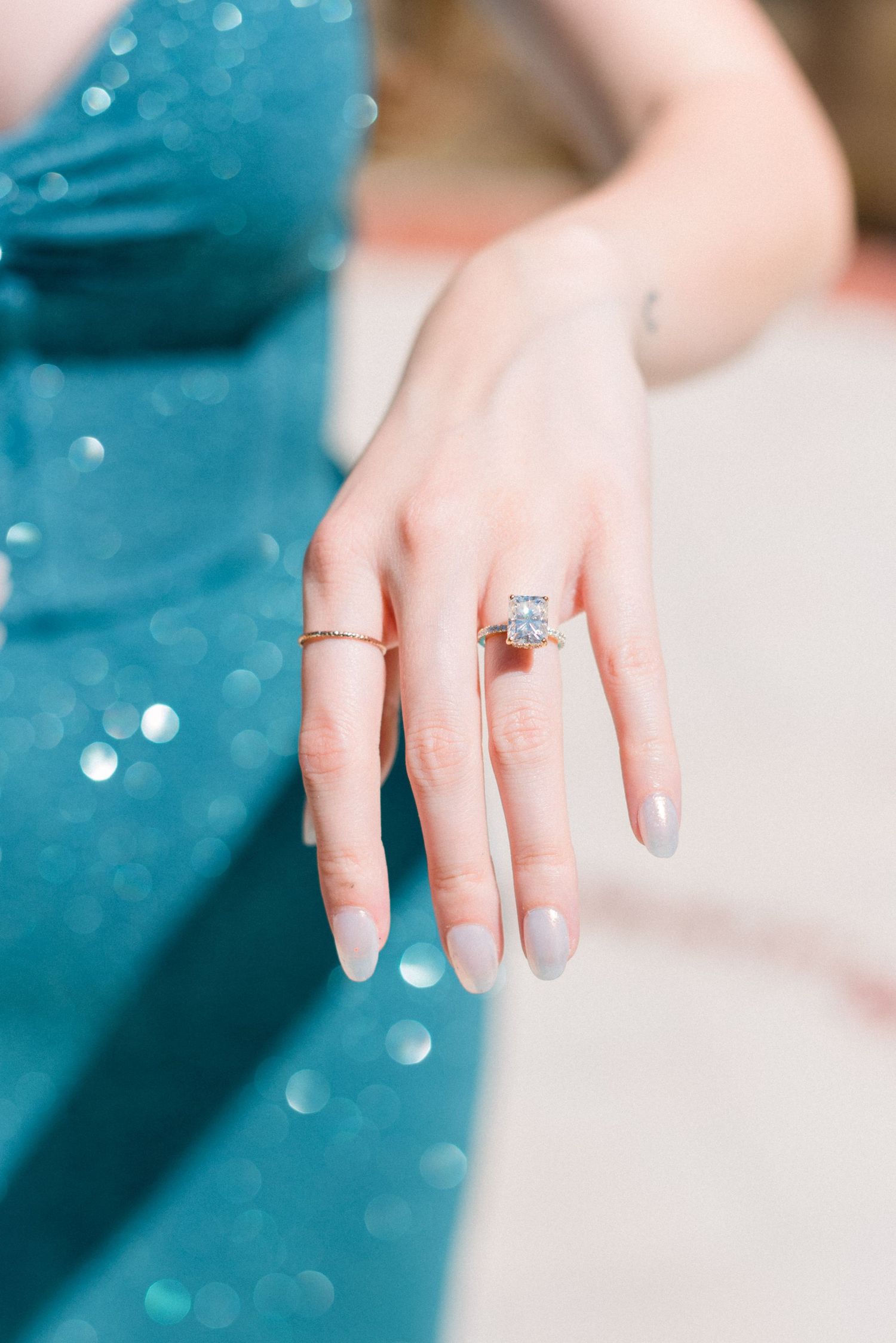 Close up of an engagement ring on a hand with a sparkly teal dress in the background.