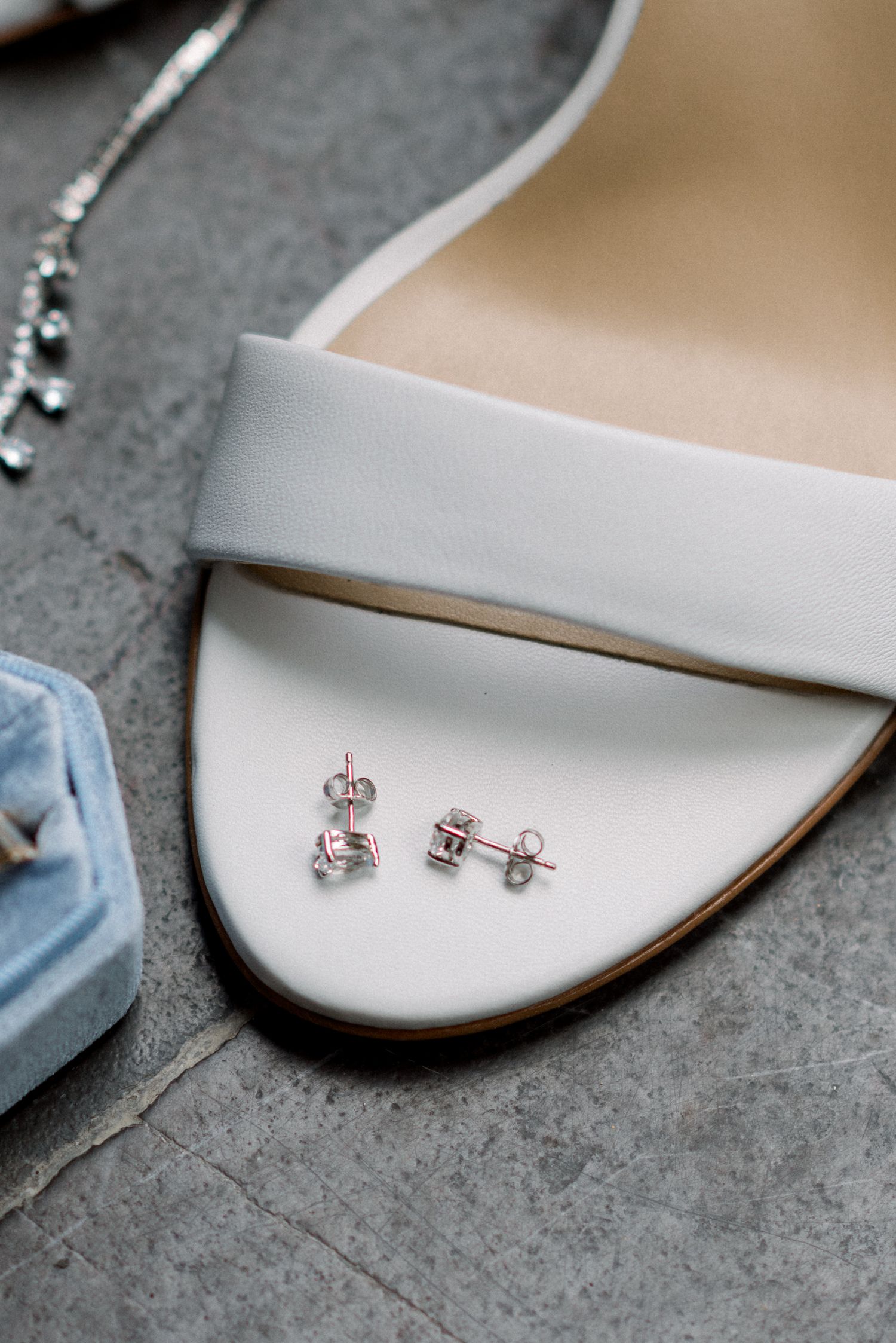 Delicate diamond stud earrings resting on a white bridal shoe against a gray background.