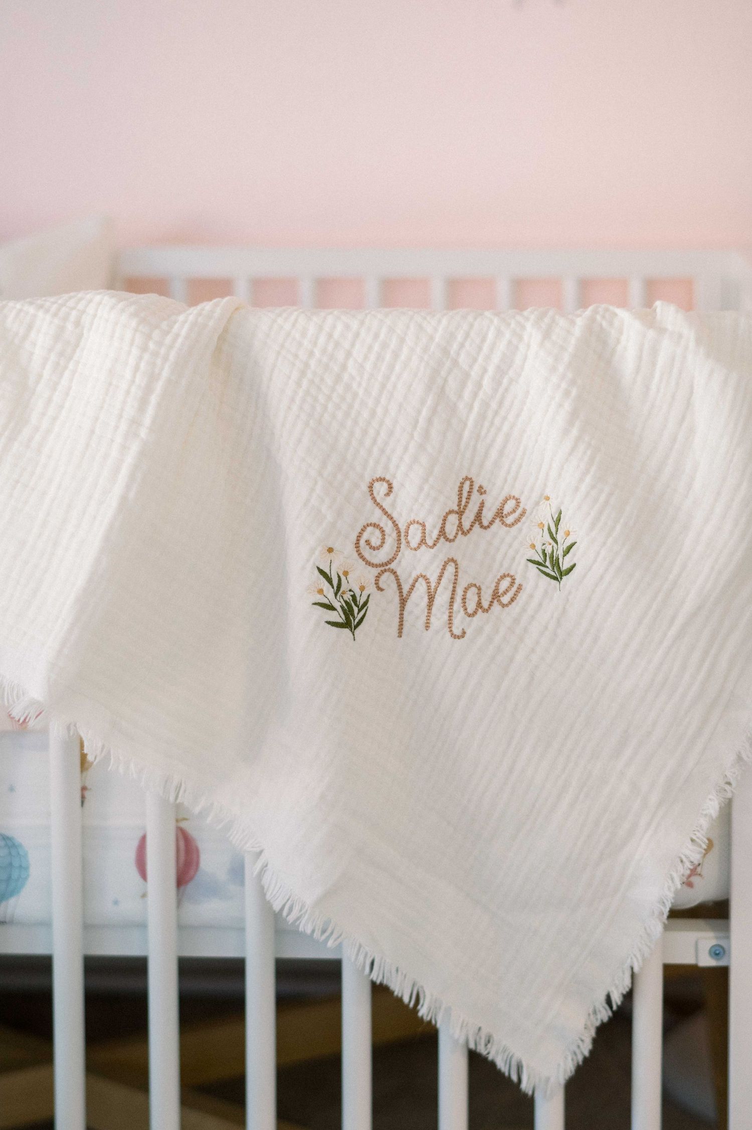 White knitted baby blanket with 'Sadie Mae' embroidered in cursive and green leaves on a crib.