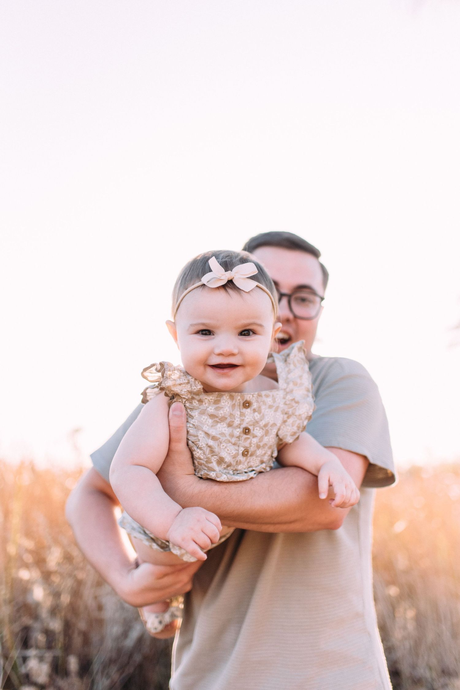 A joyful baby wearing a bow headband and floral dress is held up by someone in an outdoor setting at sunset.