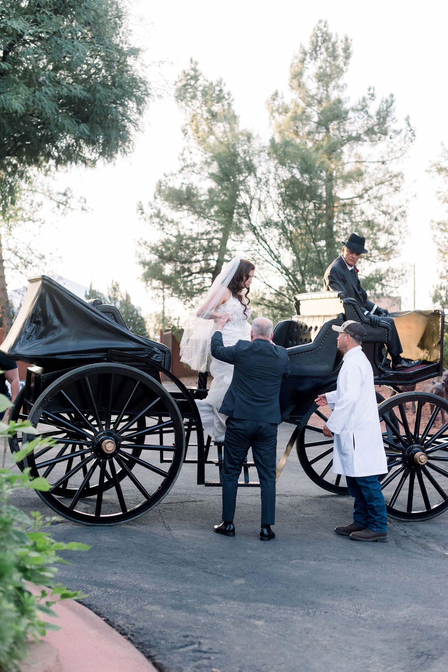 Attendants help with boarding a classic black horse-drawn carriage on a garden path.