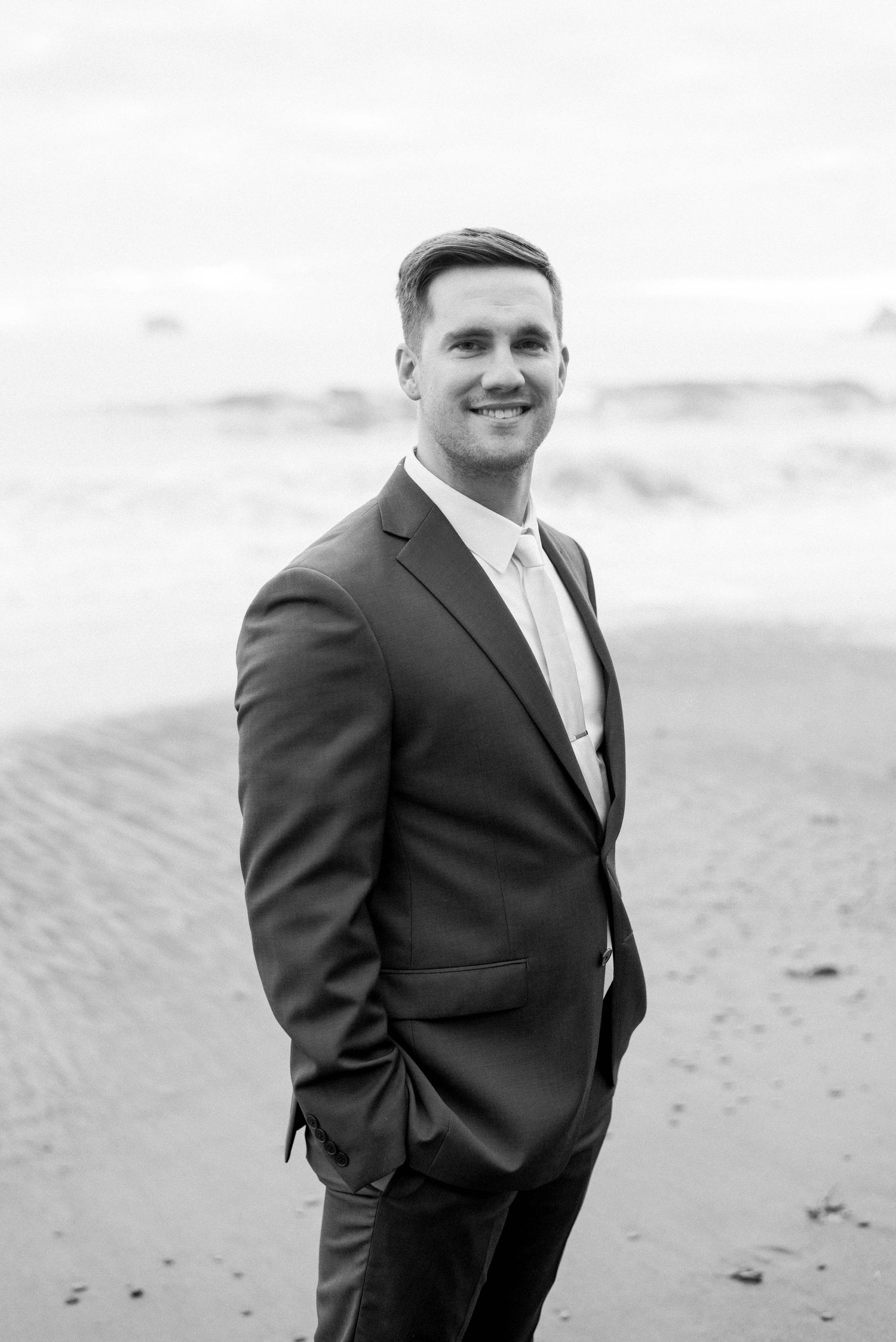 Portrait of a person in a dark suit smiling on a beach in black and white.