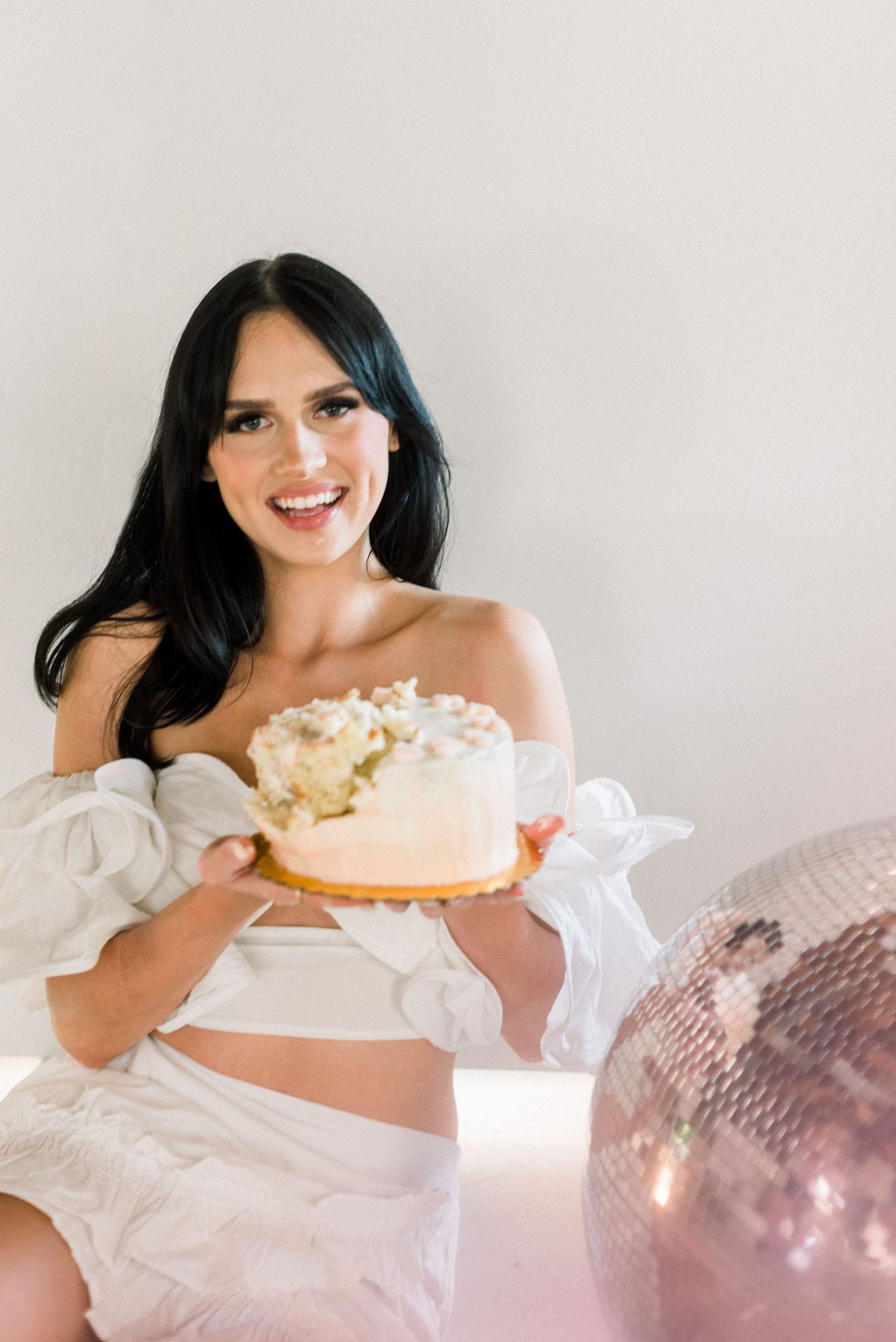 Person in white outfit holding decorated cake with disco ball in background.