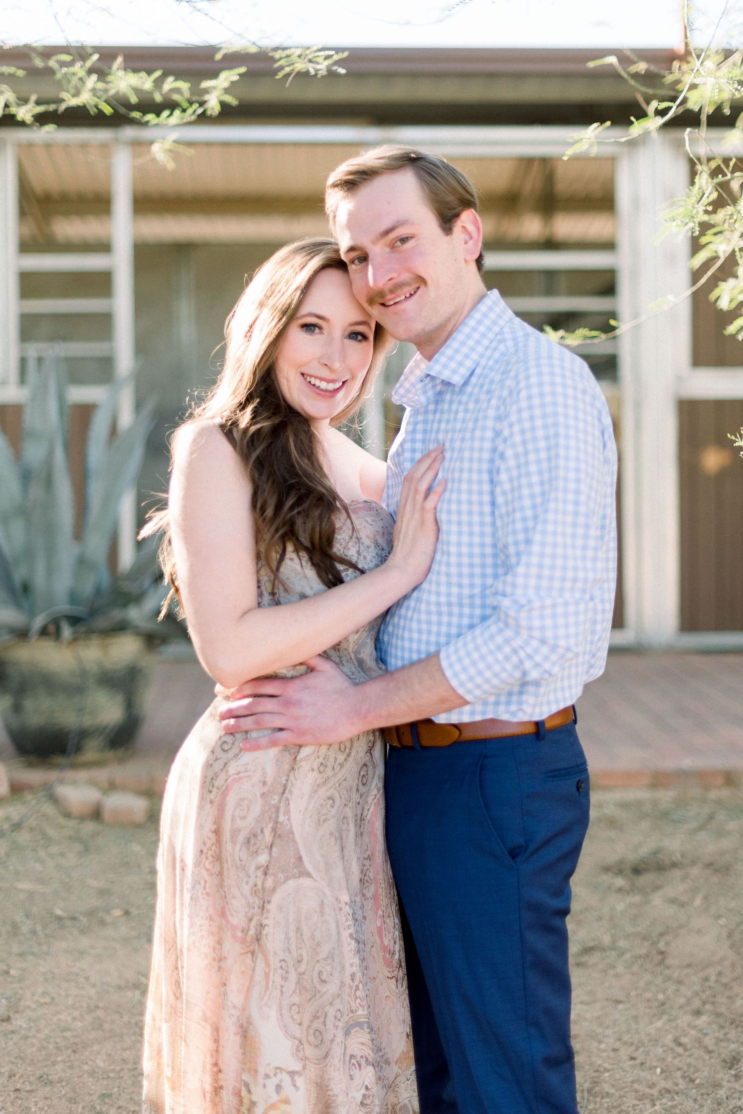 Couple embraces outdoors in romantic engagement photo with woman in lace dress and man in checkered shirt.