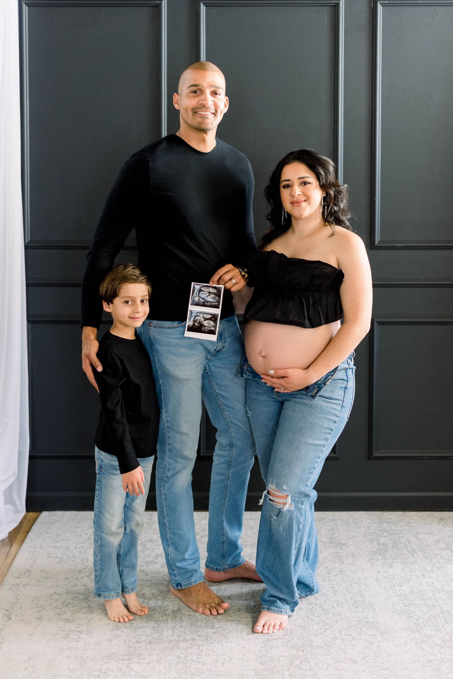 Family maternity photo with couple and child holding ultrasound photo against dark wall.