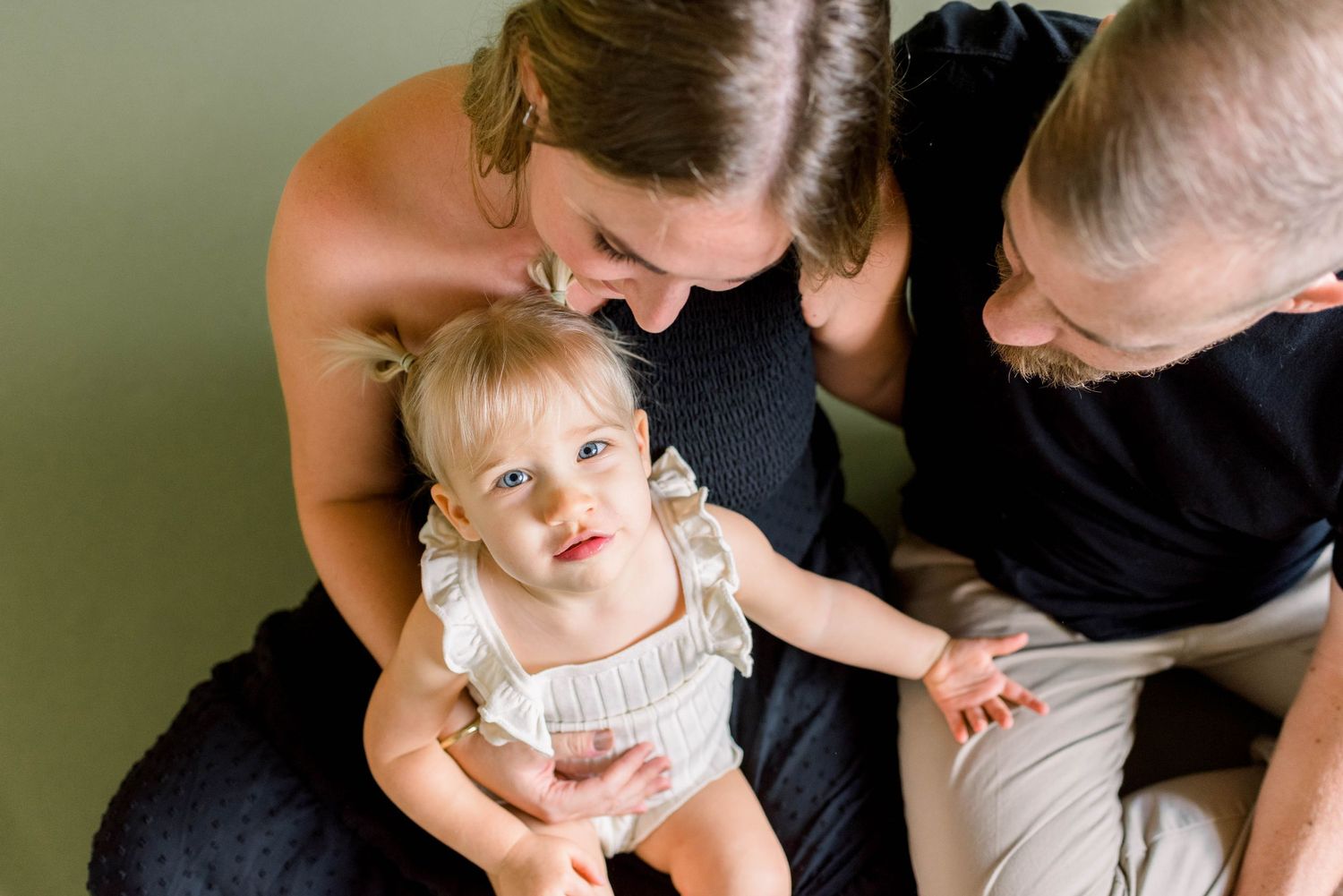 Parents lovingly embrace their young toddler in a candid indoor family portrait against a neutral background.