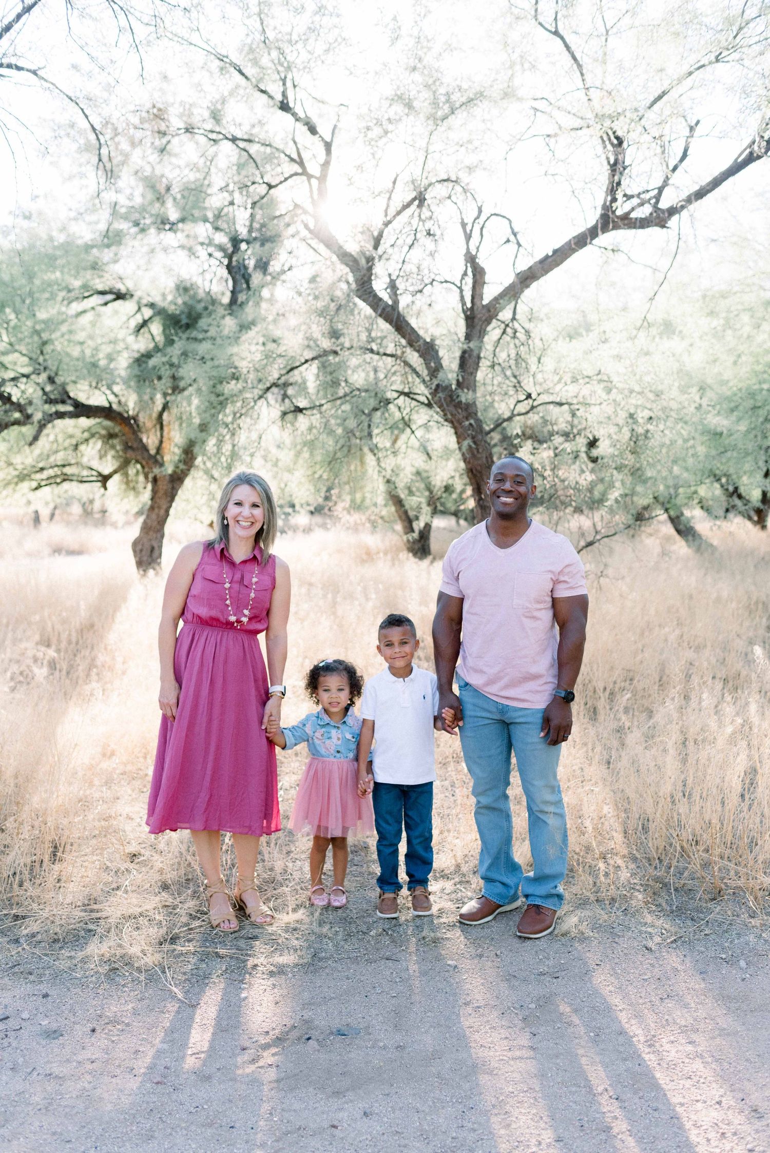 A family stands together in casual attire under olive trees casting long shadows on a dusty path.