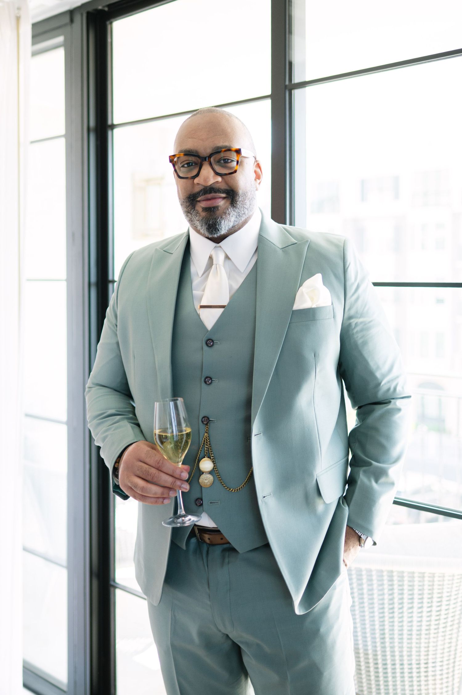 A person in a light blue suit and glasses poses confidently near large windows.