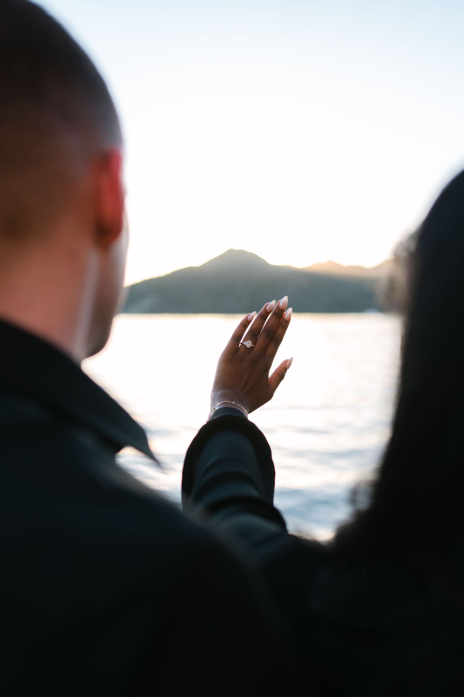 Silhouetted figures gesturing against a bright sky with water and mountains visible in the background.