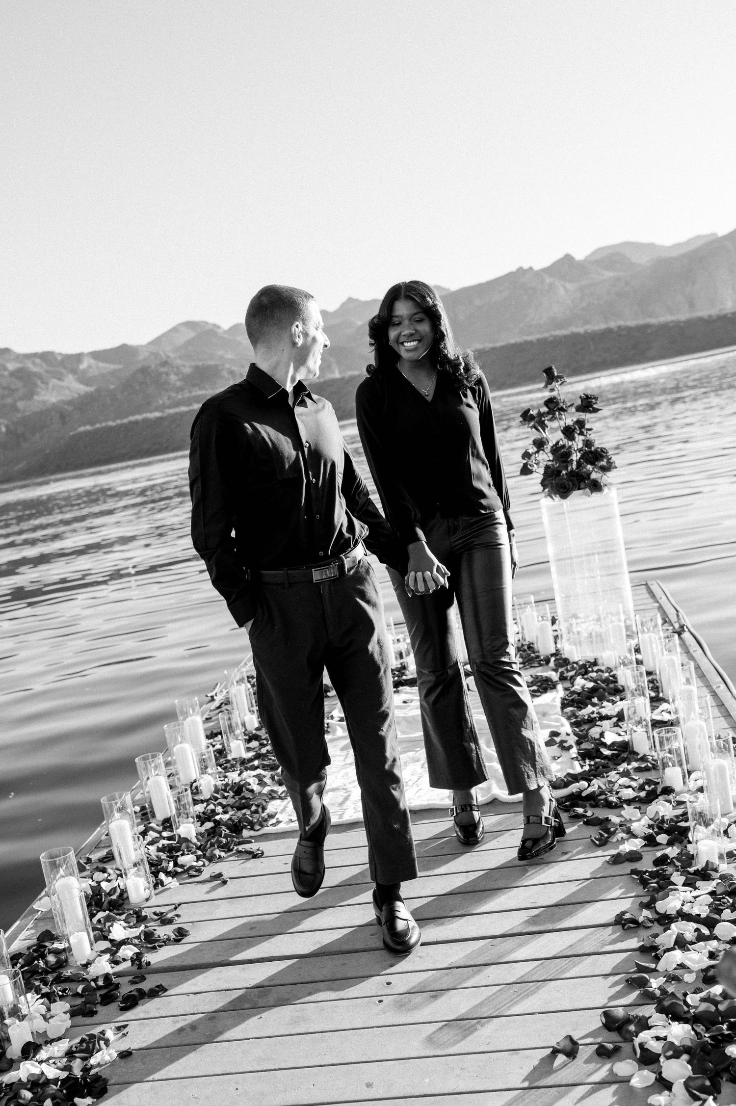 Black and white photo of a couple walking along a waterfront deck decorated with floral arrangements and mountains behind.