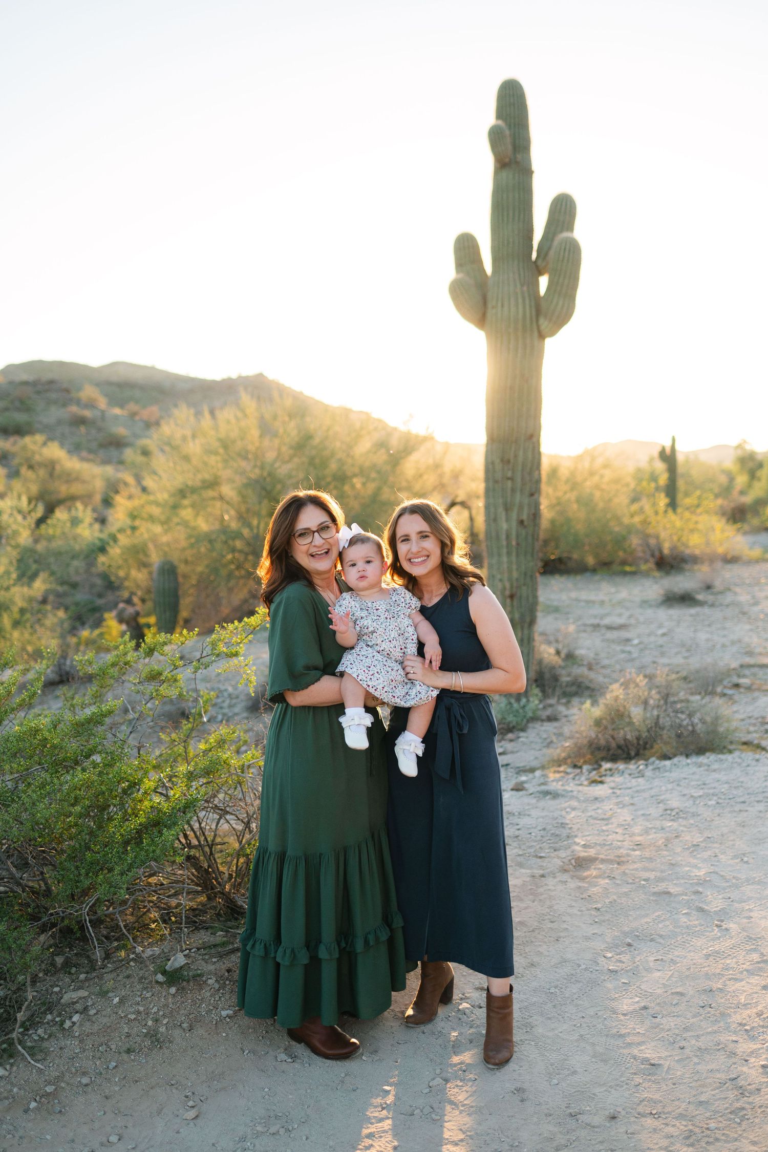Family in coordinated green and navy outfits poses together in desert landscape with saguaro cactus at sunset.