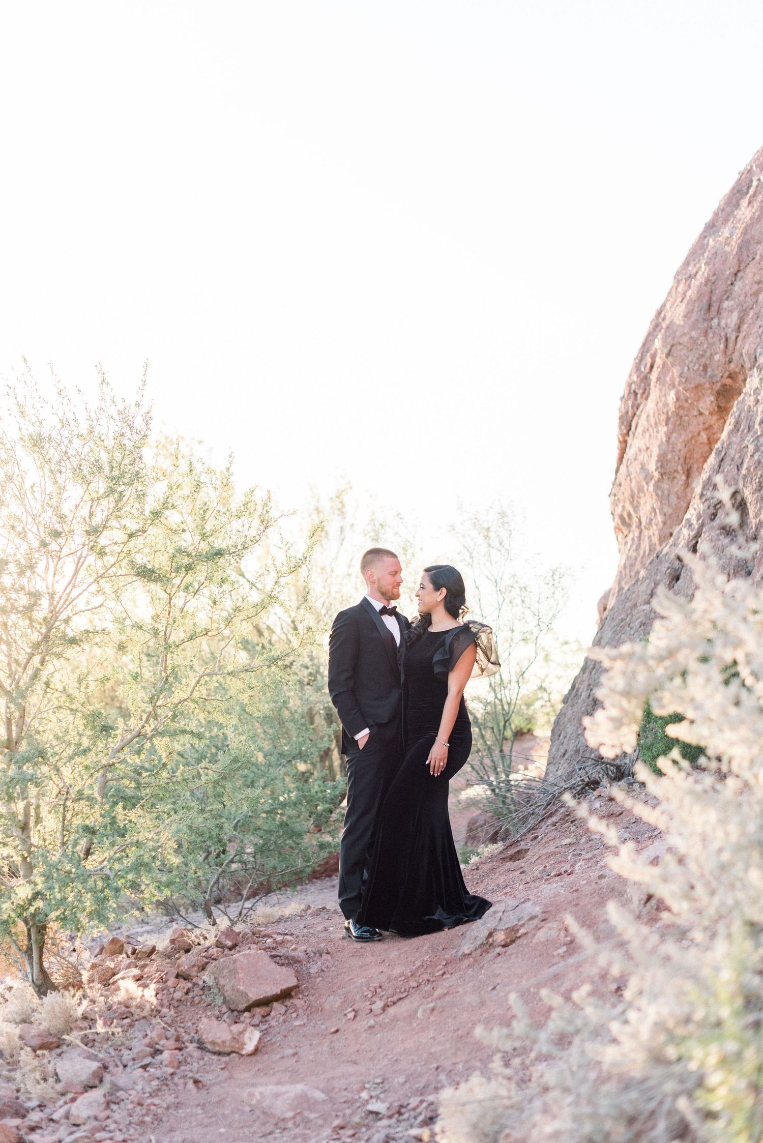 Couple in formal attire sharing an intimate moment while sitting on a rocky desert trail during sunset.