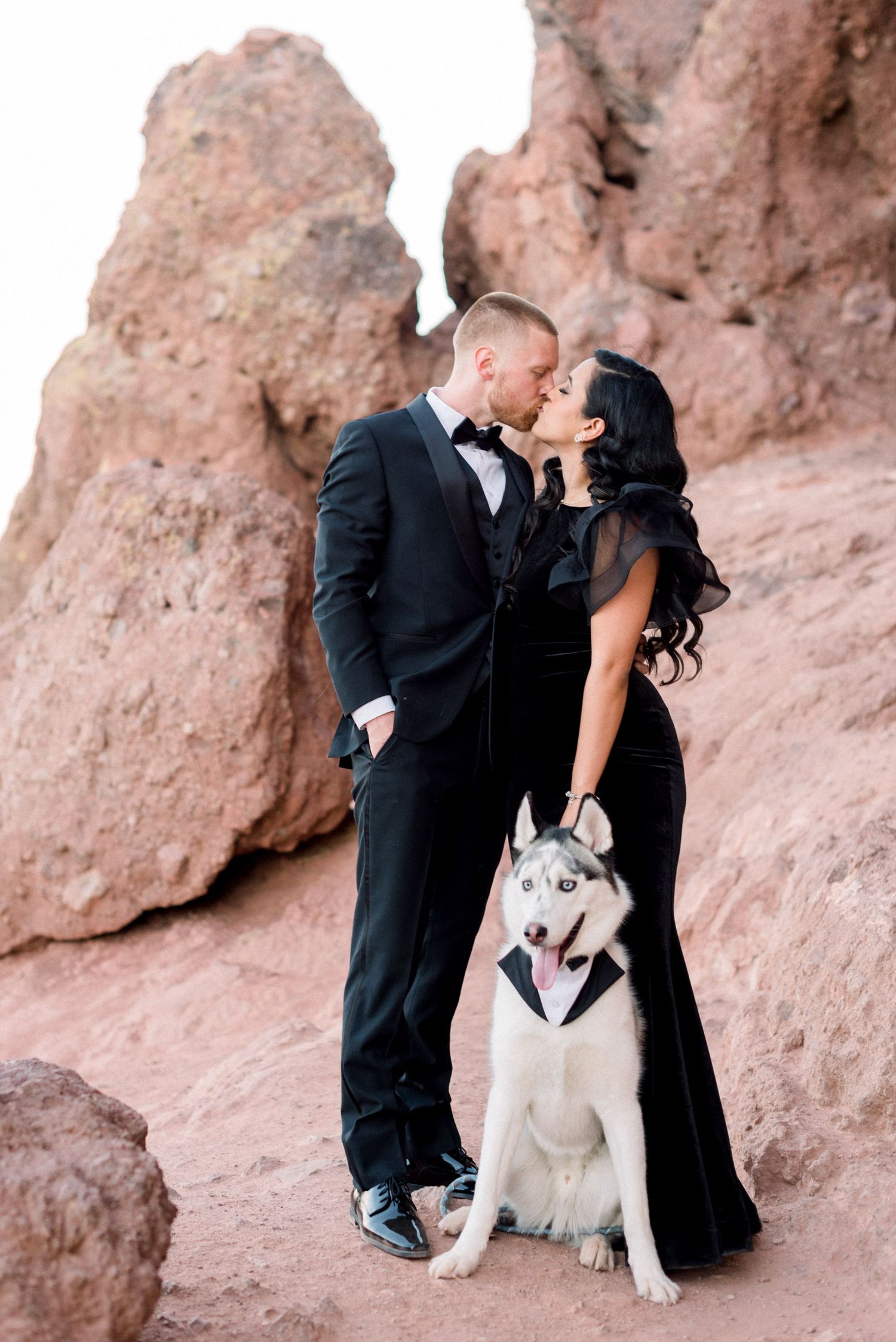Elegant couple in black attire poses with their husky dog against red rock formations.