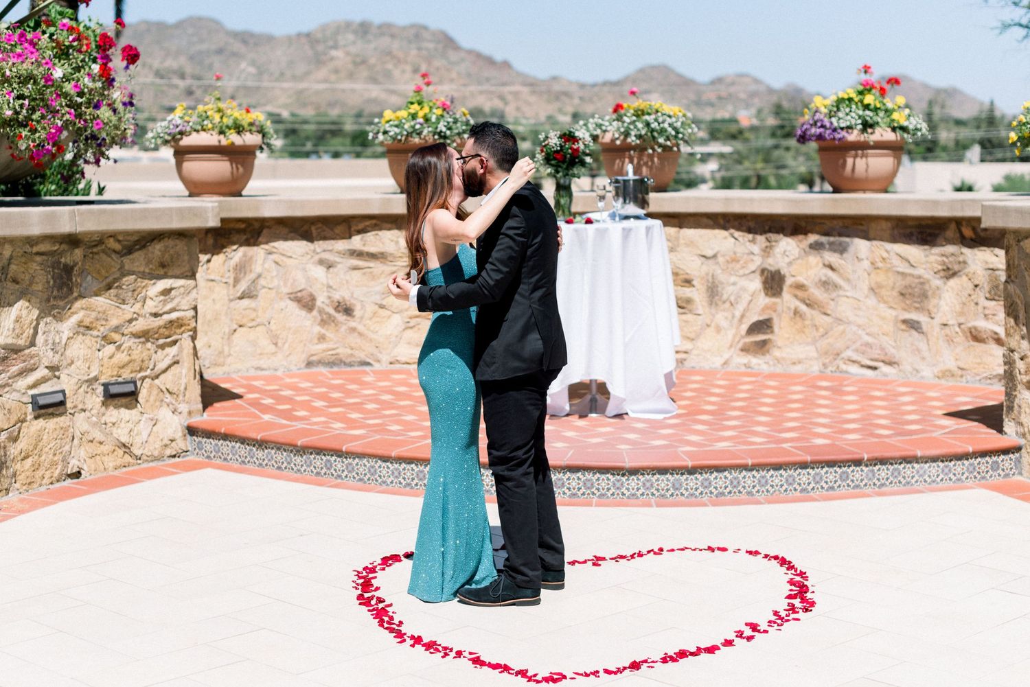 Newly engaged couple embracing inside a heart made of rose petals on an outdoor terrace.