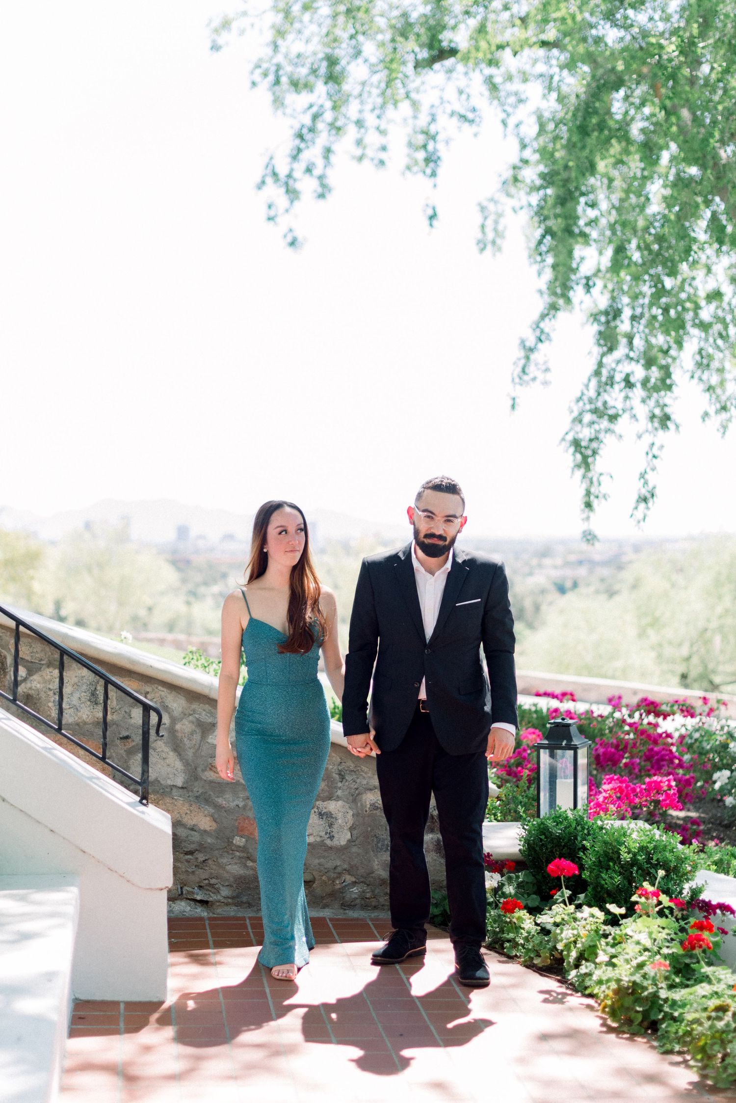 Couple walks hand in hand on a garden path with blooming flowers and greenery.
