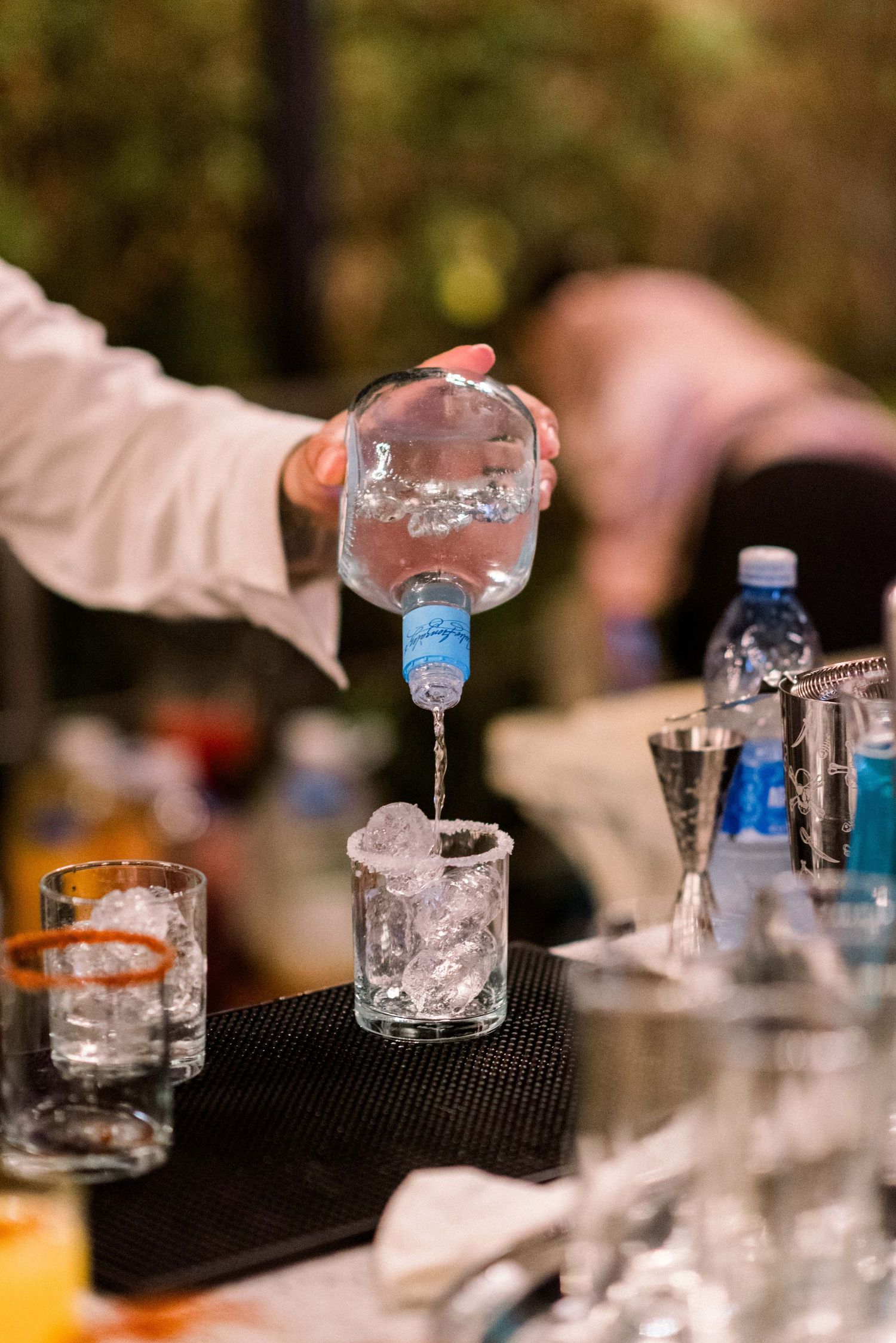 Bartender pouring clear liquid into ice-filled glass at outdoor event.