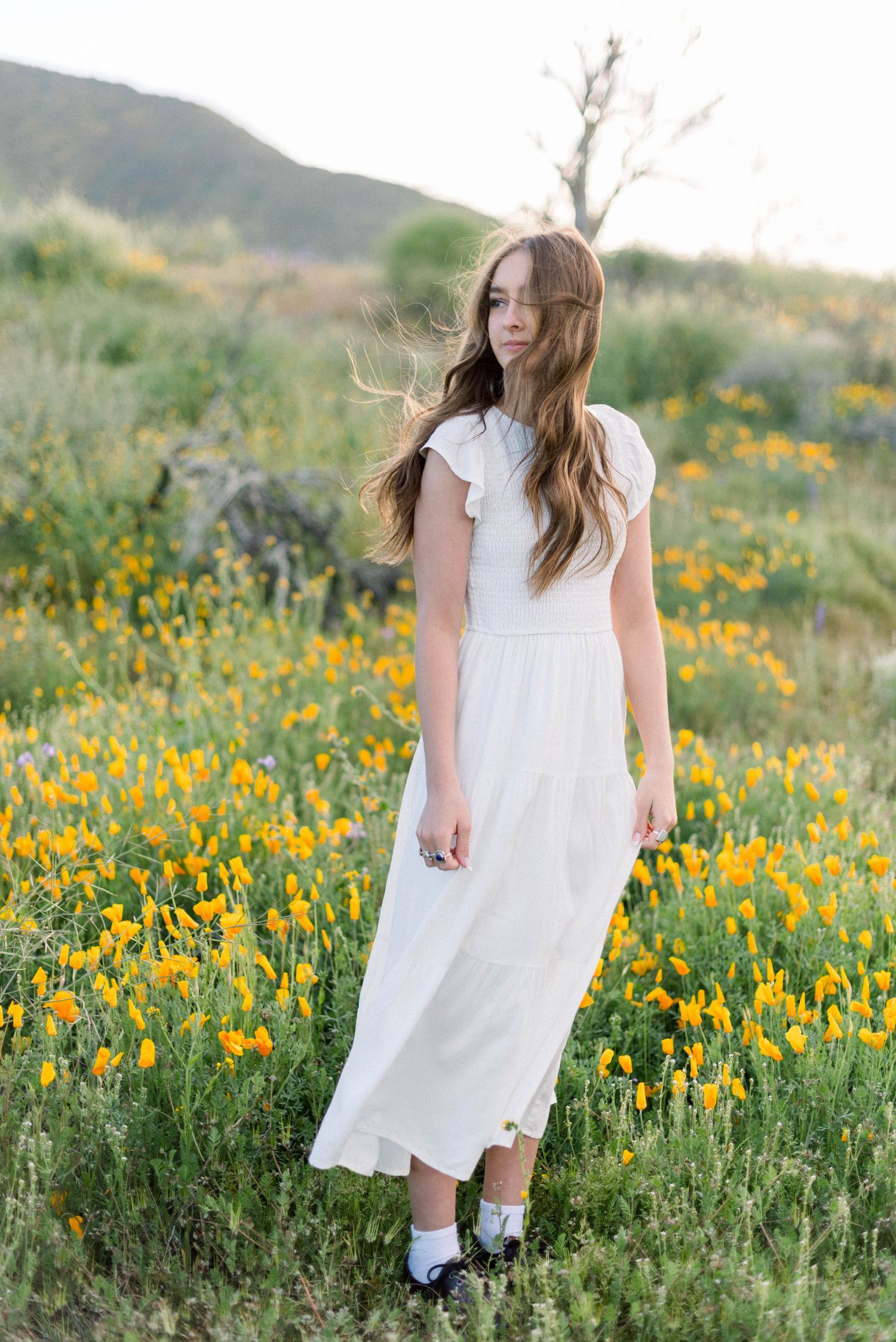 Long white dress stands gracefully among field of yellow wildflowers with mountains behind.