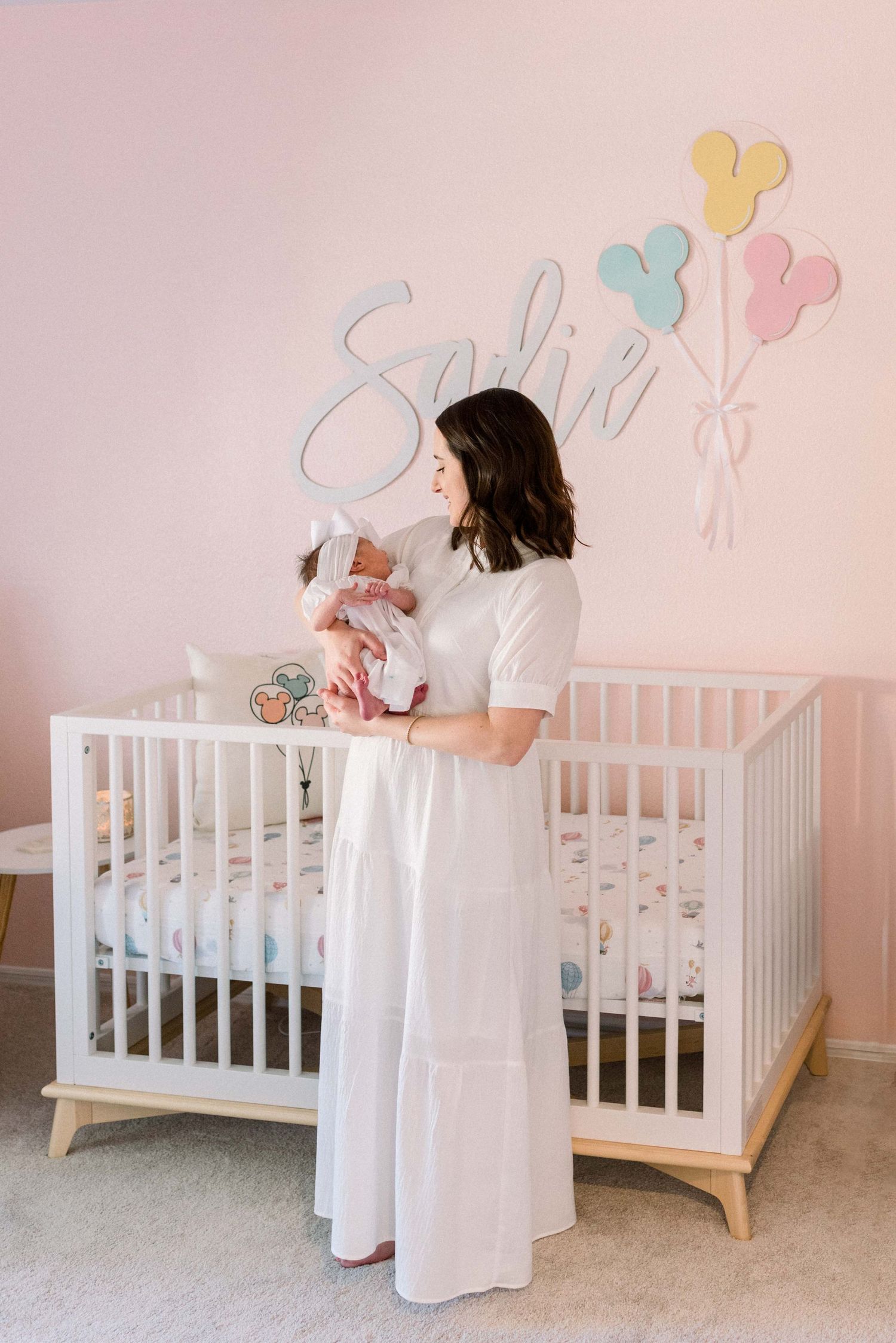 Woman in white dress holding baby in front of Disney-themed nursery wall.