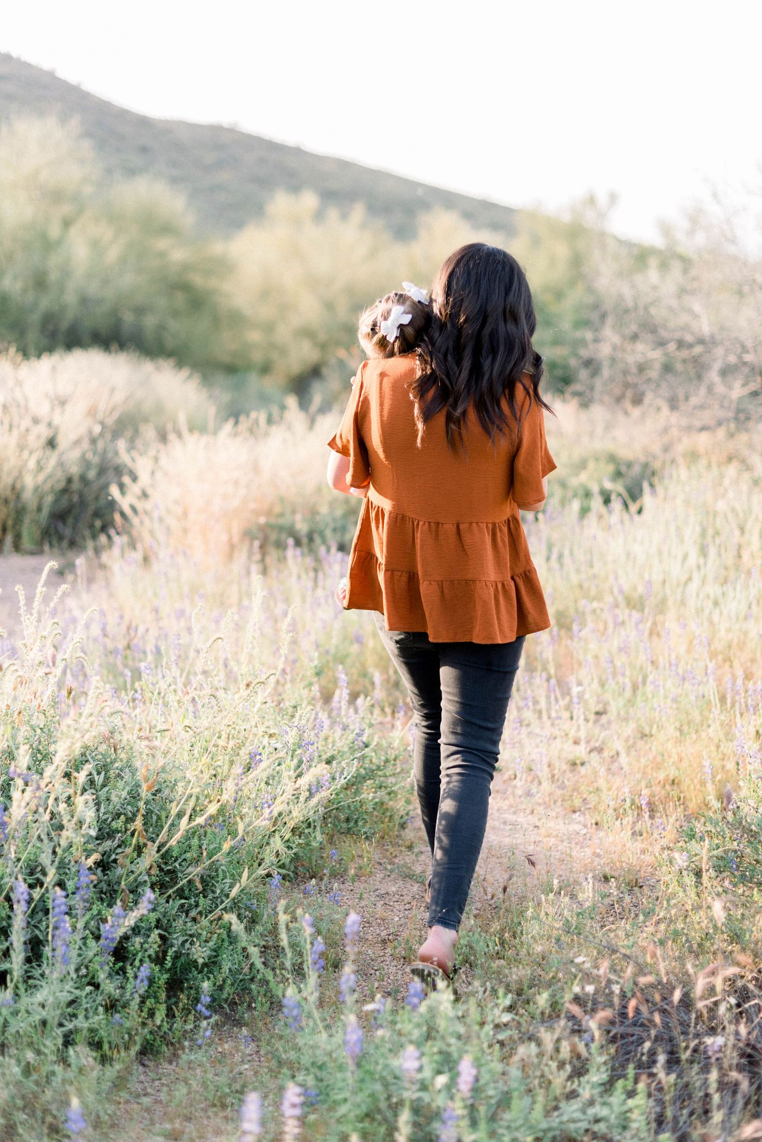 Person in rust-colored top walking through desert wildflowers with mountains in background.