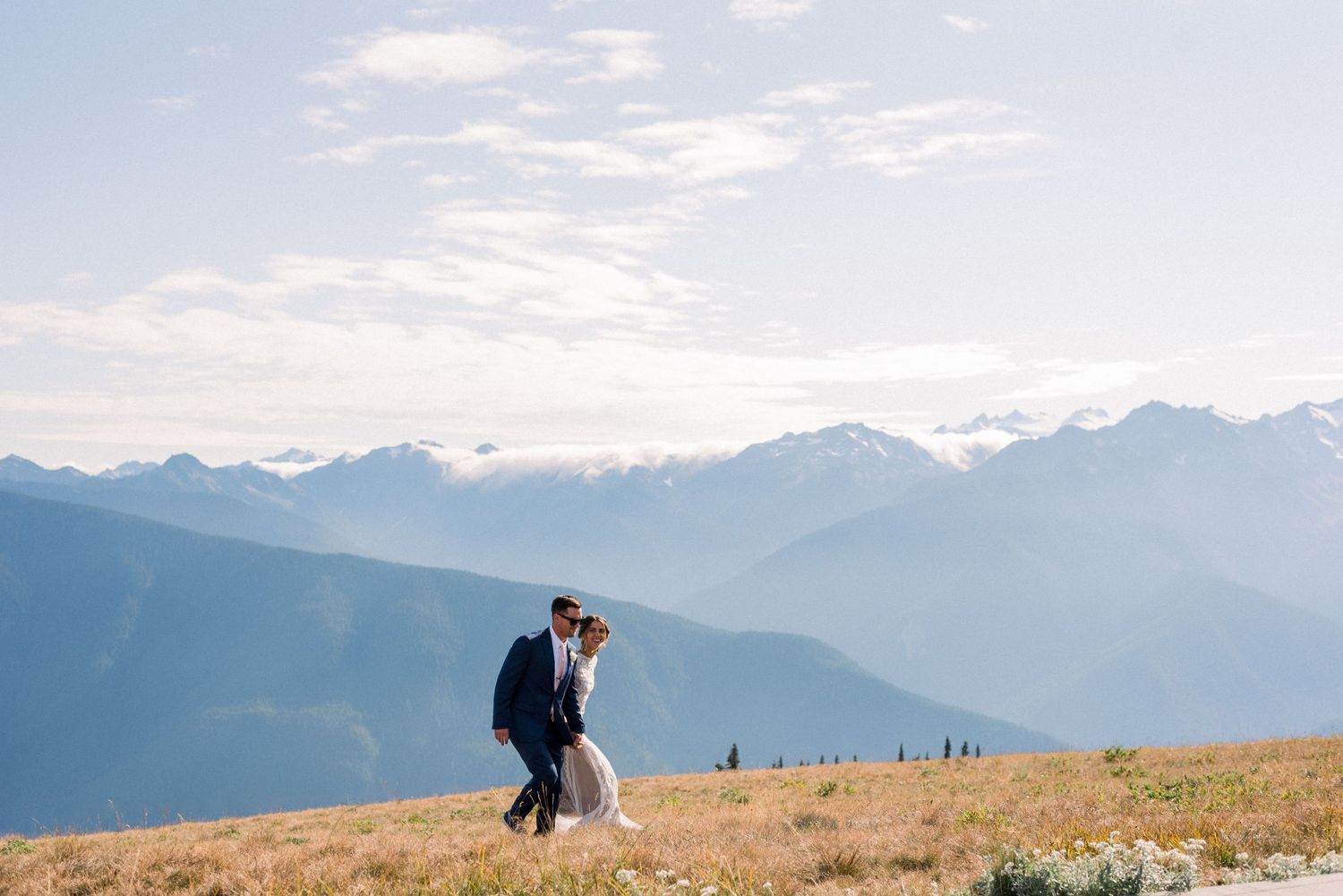 Wedding couple walks hand in hand across an alpine meadow with mountain peaks in the distance.