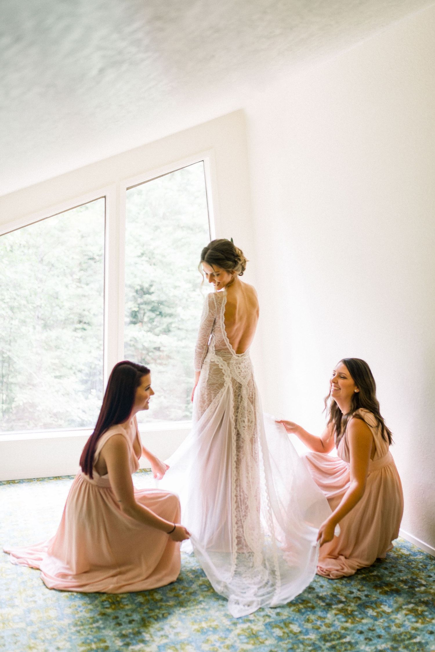 Bridesmaids helping with wedding dress preparations in a bright room with large windows.