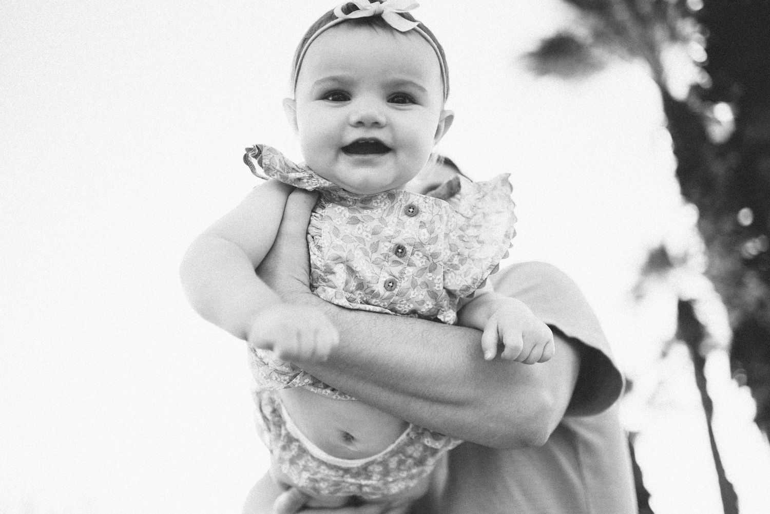 Black and white portrait of a happy baby in a ruffled dress being held up outdoors.