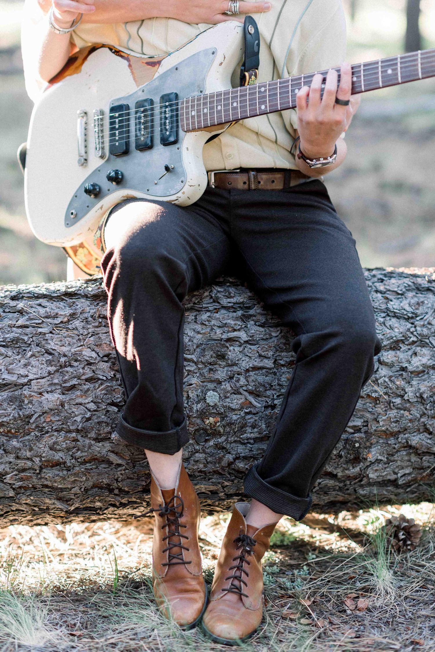 Close-up of brown boots and a white electric guitar while sitting on a fallen log in the woods.