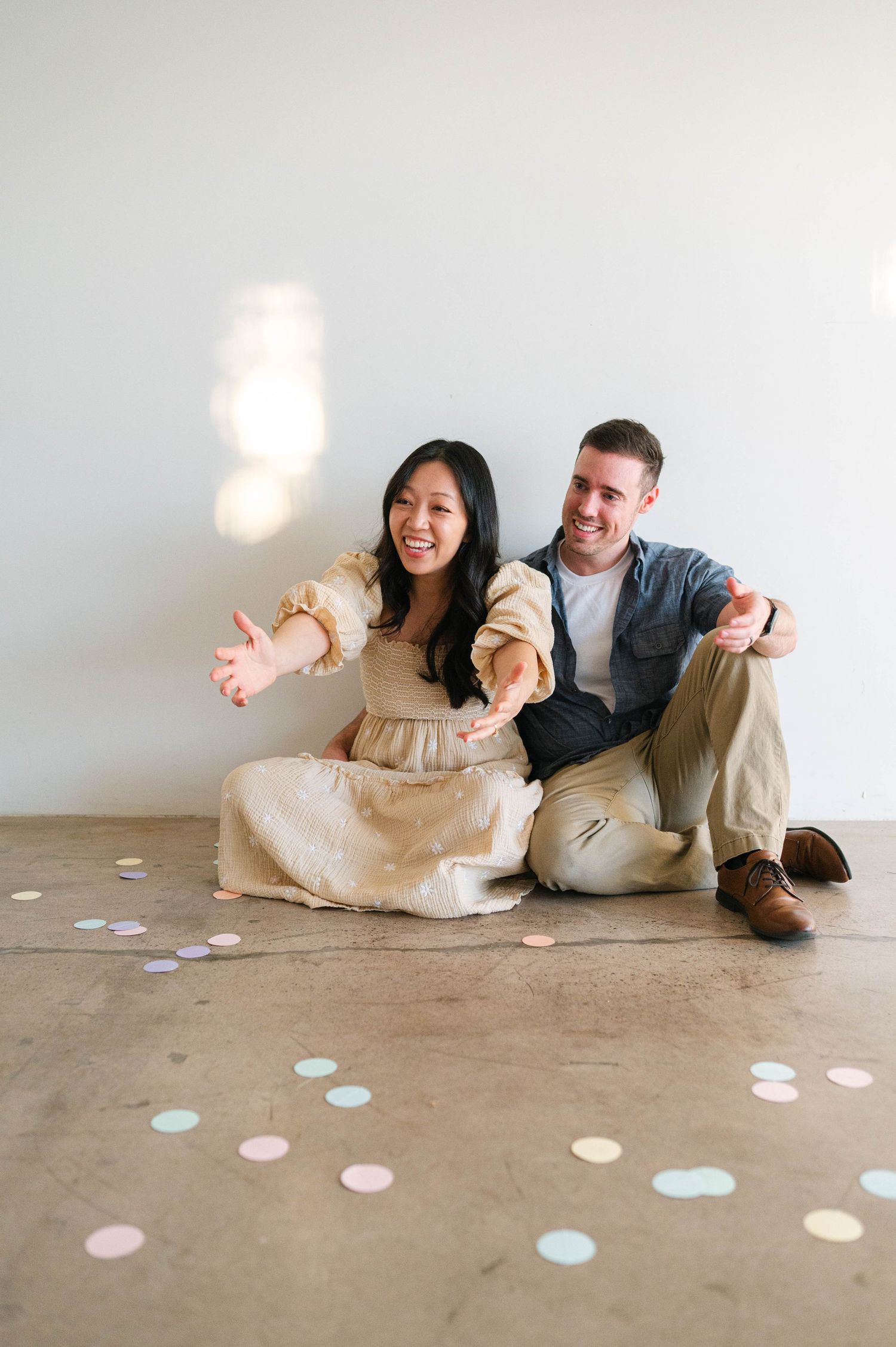 A couple sits on the floor surrounded by pastel confetti dots in a minimalist setting.