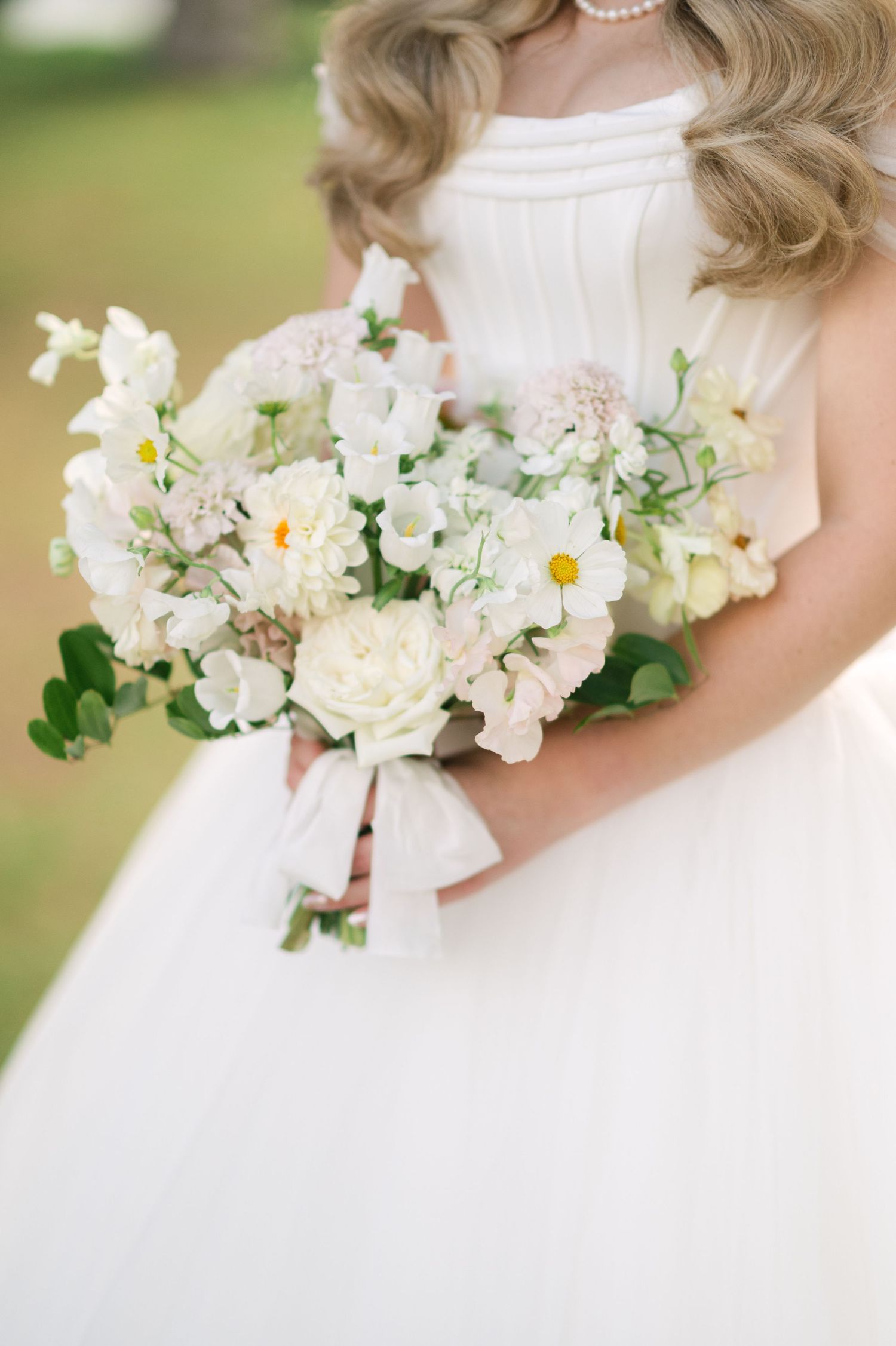 Close-up of elegant white wedding bouquet with delicate white flowers and greenery against a light background.