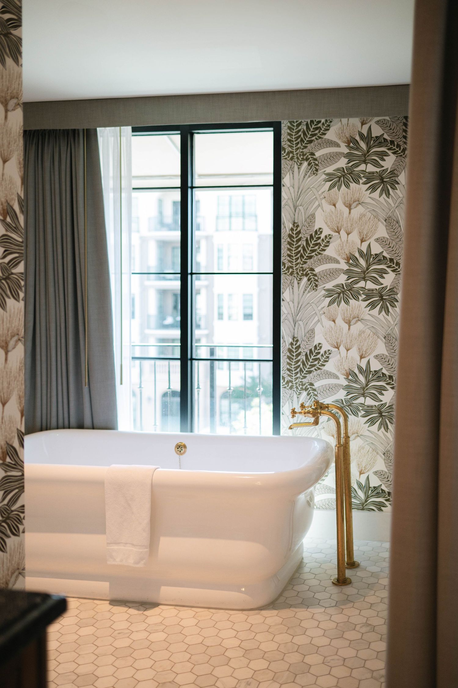 Luxurious modern bathroom featuring a freestanding white tub, gold faucet, floral wallpaper and hexagonal floor tiles.