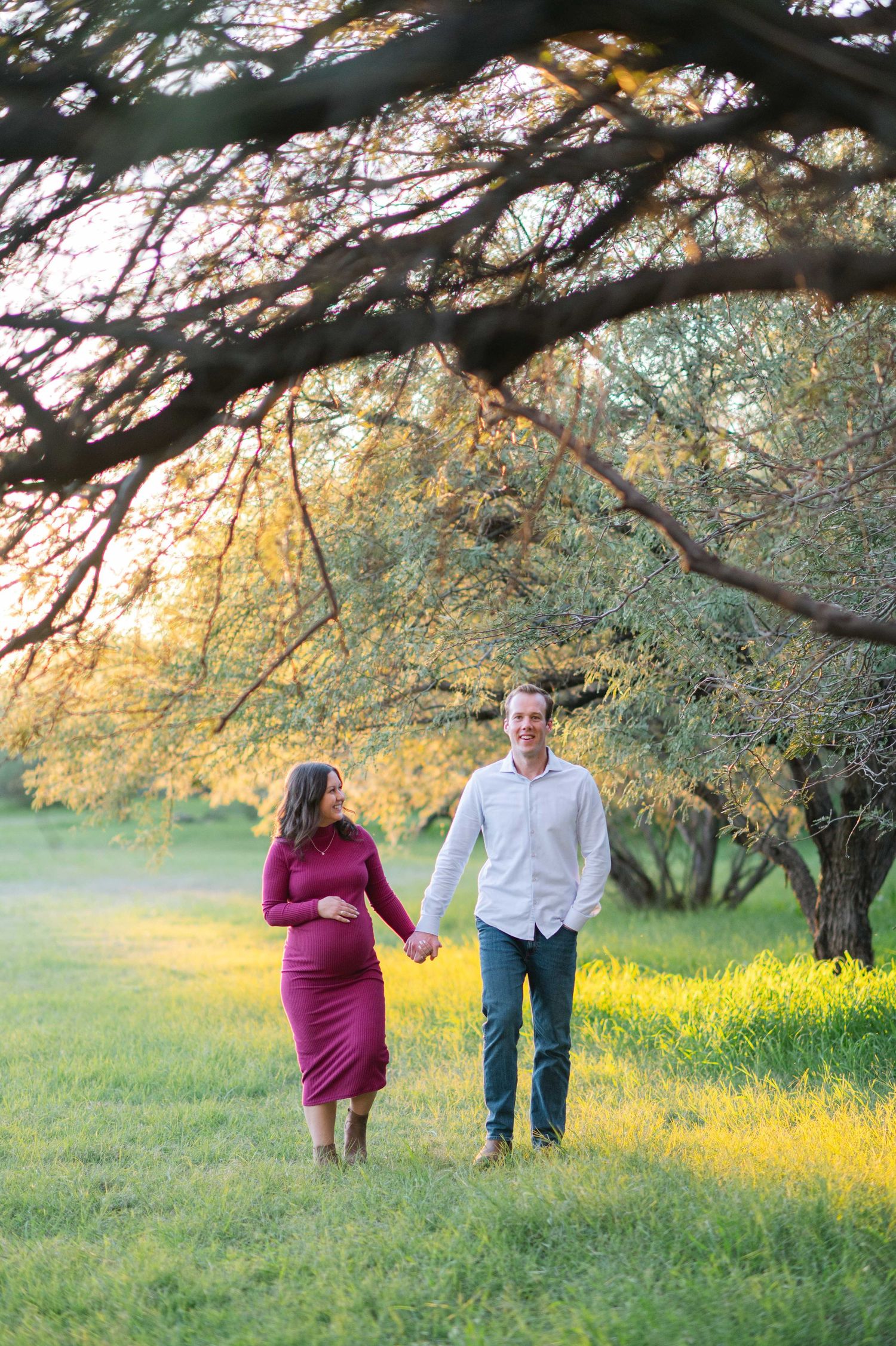 A couple walks hand in hand through a meadow under blooming spring trees at sunset.