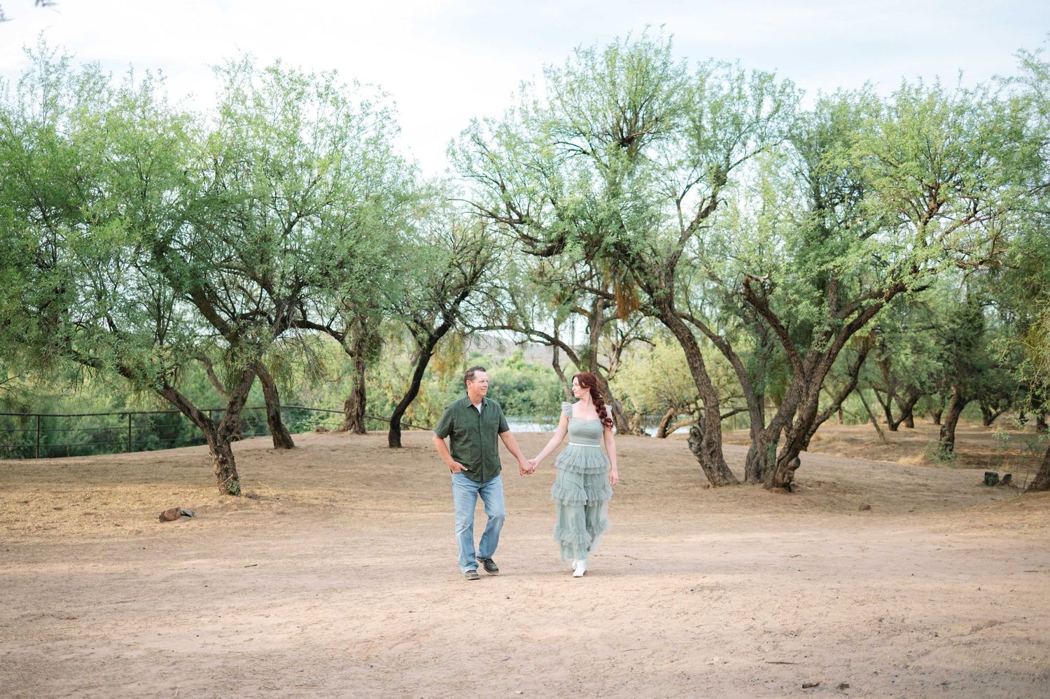 Two people walk through a grove of olive trees on a sunny day in a Mediterranean landscape.