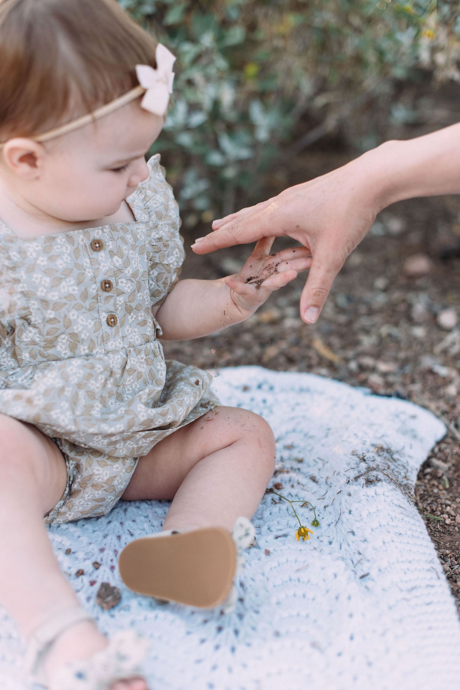 Baby in floral dress reaches for adult hand while sitting on white lace blanket outdoors.