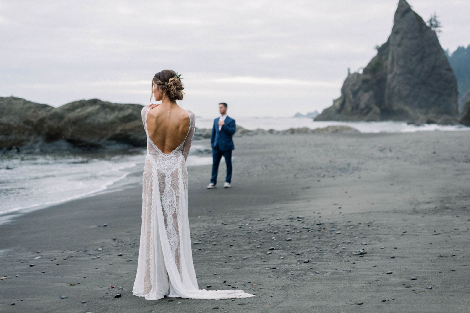 A bride in a backless gown stands on beach while groom waits in distance near coastal cliffs.