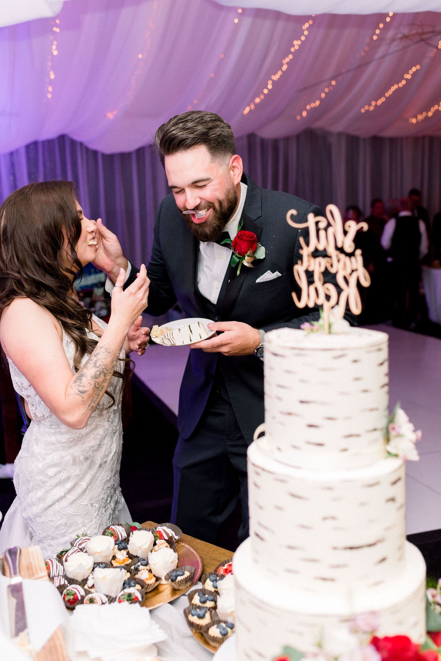 Couple shares joyful moment while feeding each other wedding cake beside elegant tiered dessert.