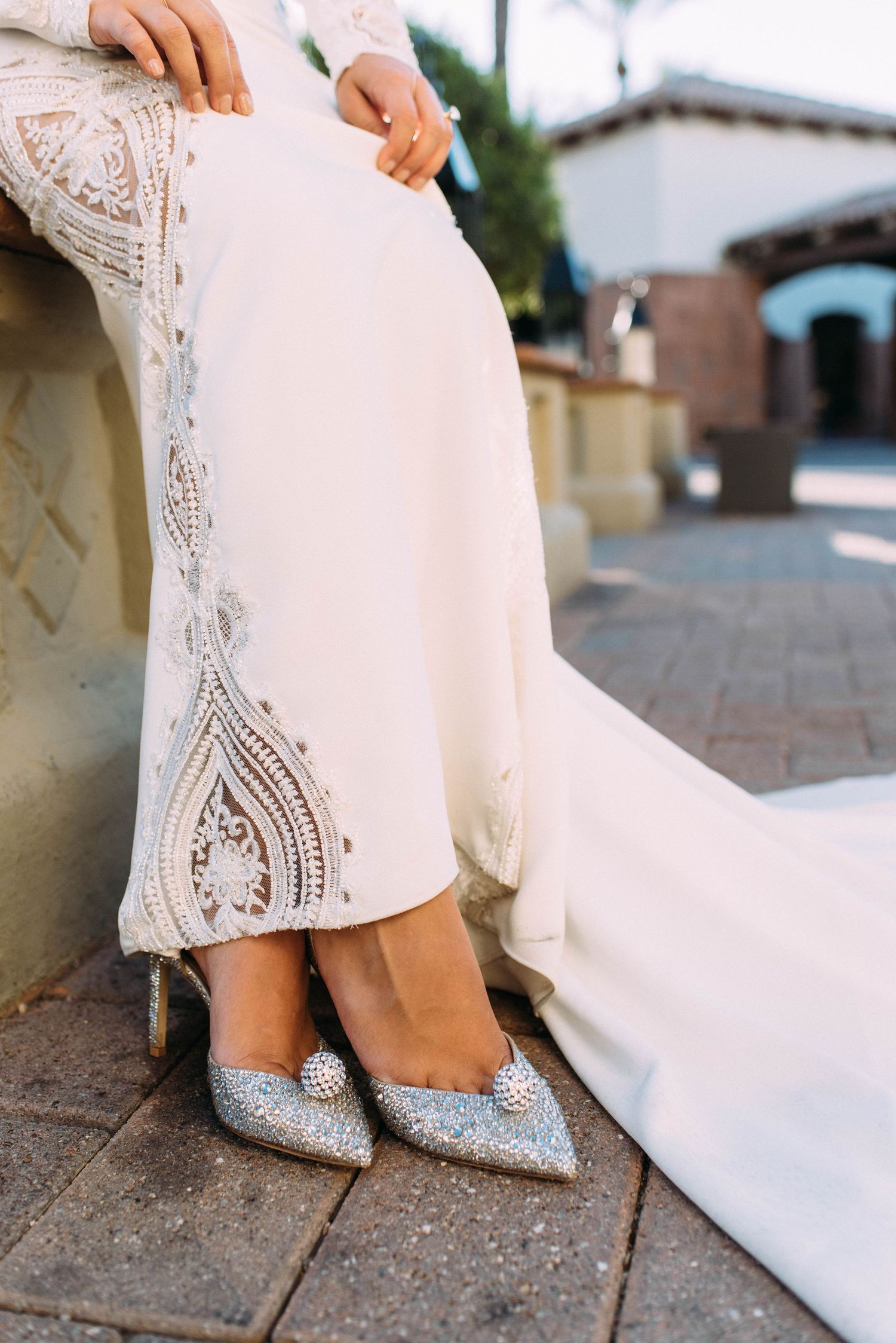 Close up of sparkly silver bridal shoes and lace dress hem details on stone pavement.