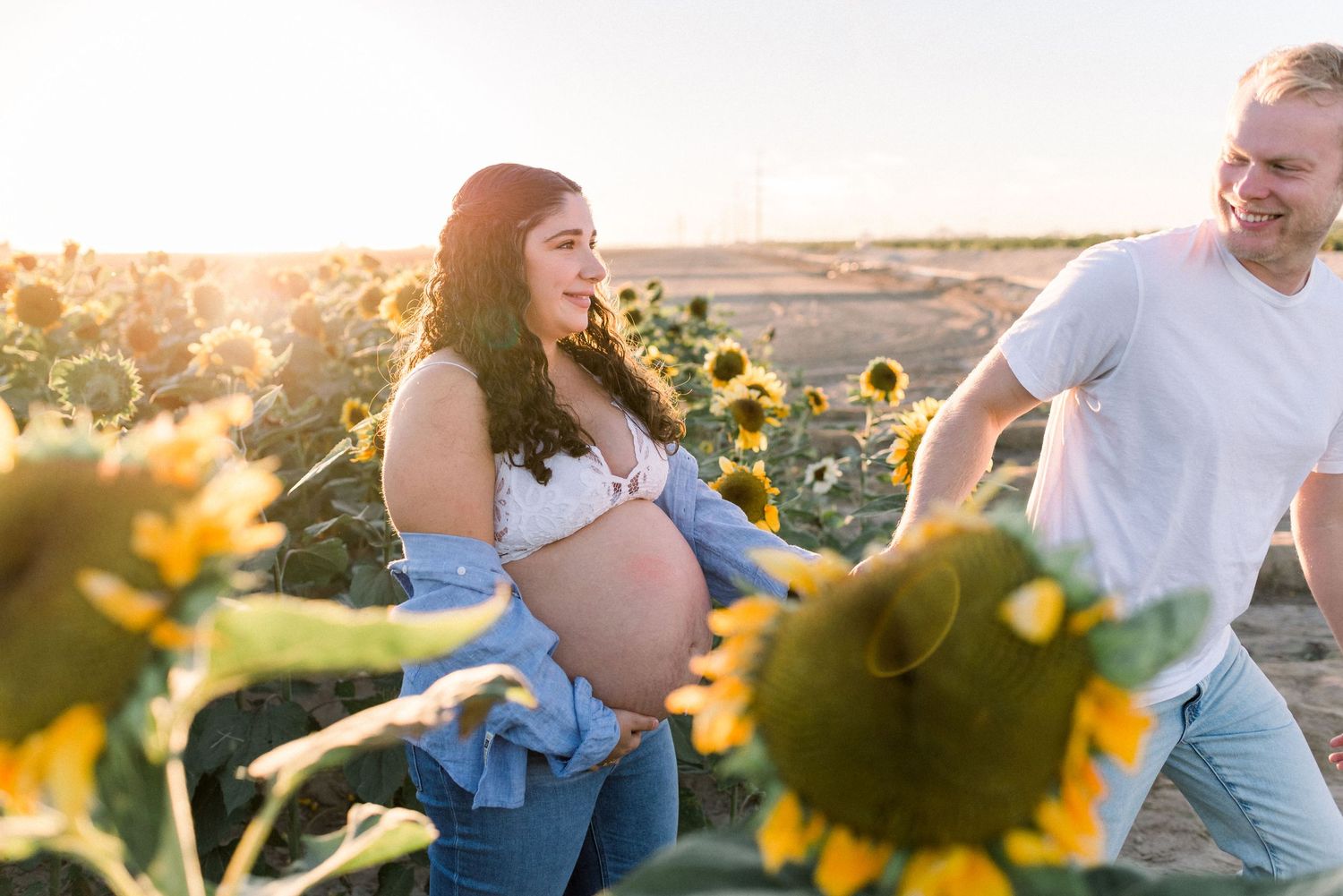 Expectant couple poses in a sunlit sunflower field during sunset for a maternity photoshoot.
