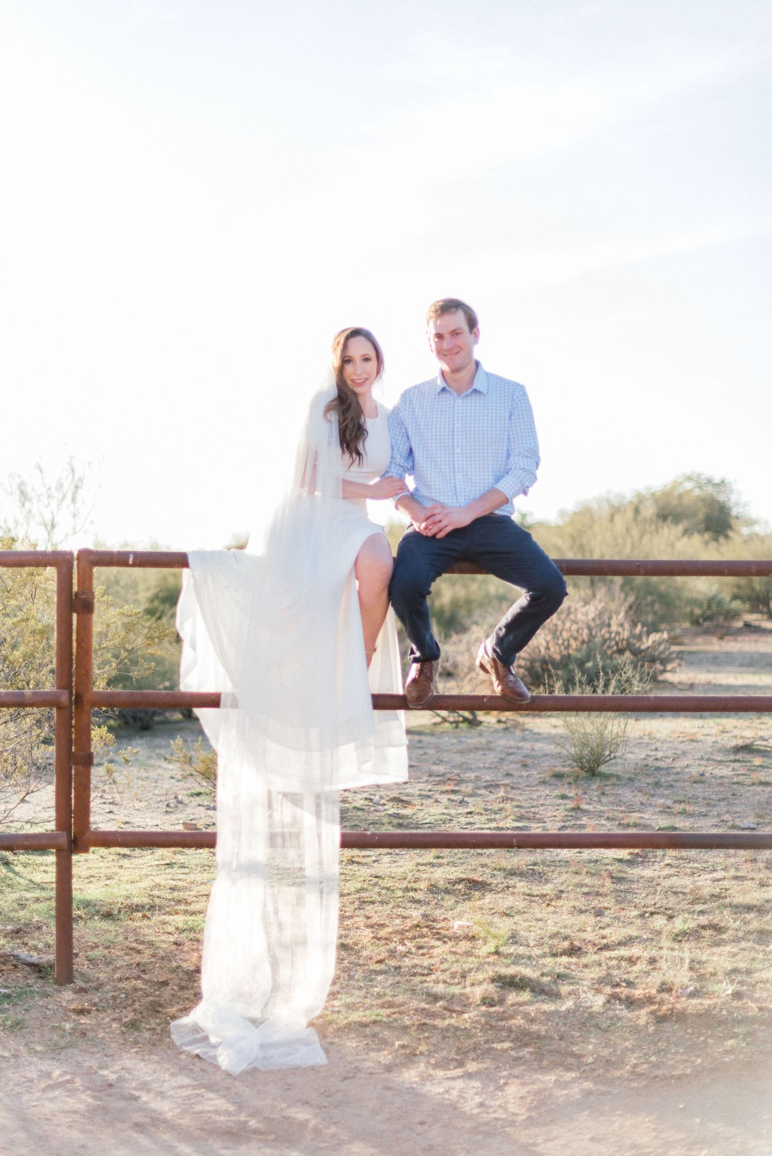 Couple in blue and white sitting on a rustic wooden fence during a desert photoshoot.