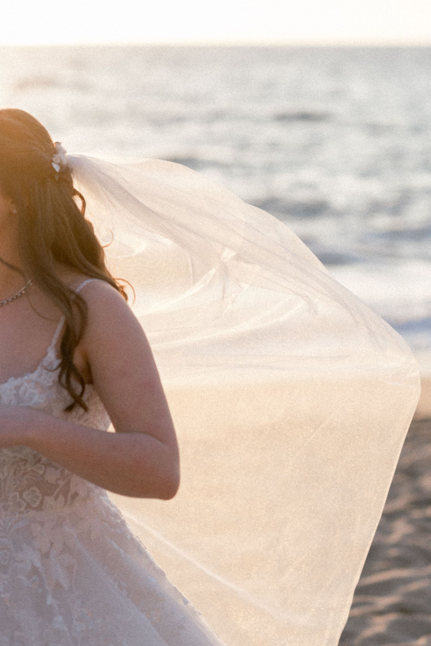 Flowing white wedding veil caught in ocean breeze on sandy beach at sunset.