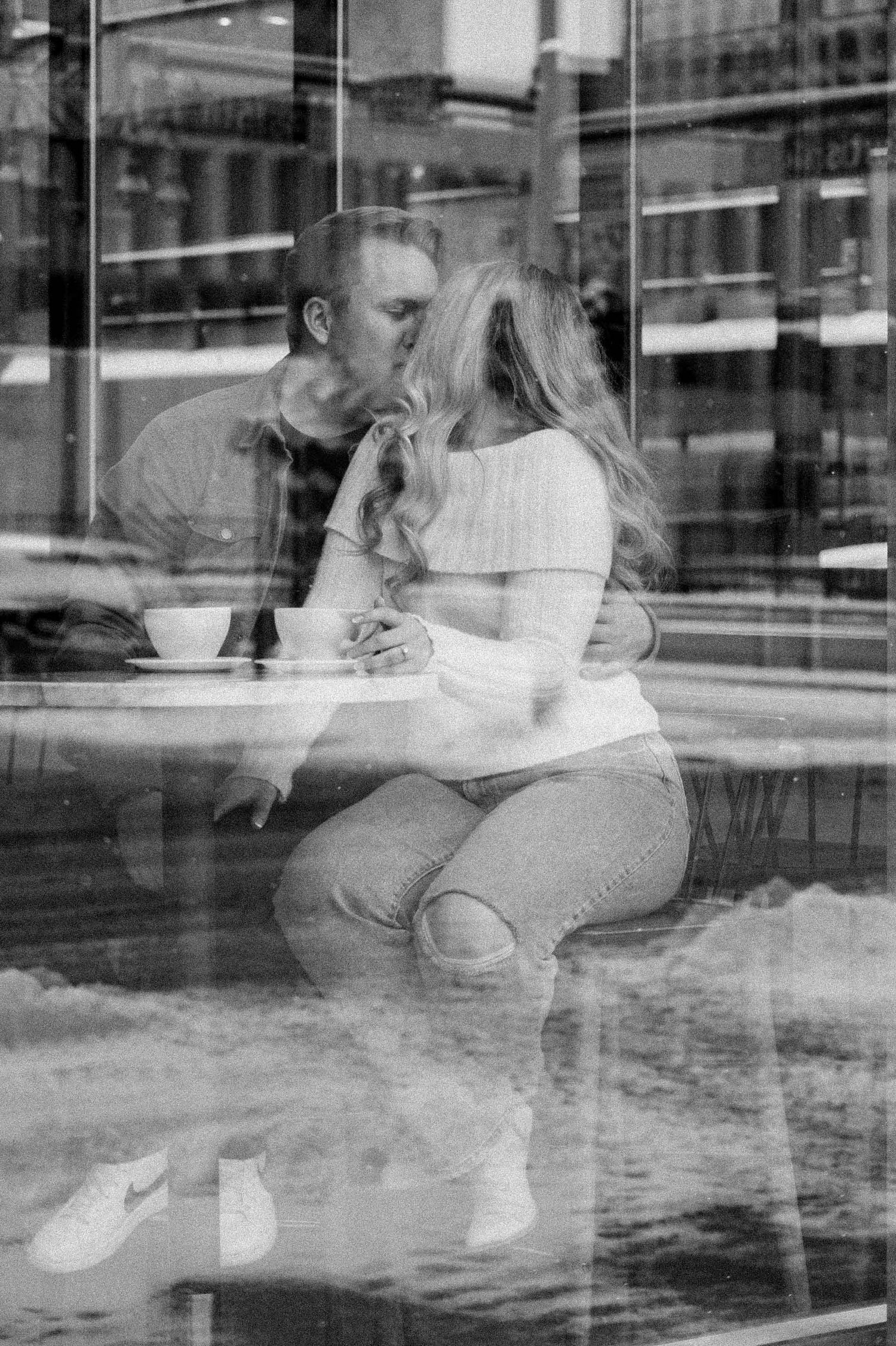 A black and white image of a couple sharing an intimate moment at a cafe window.