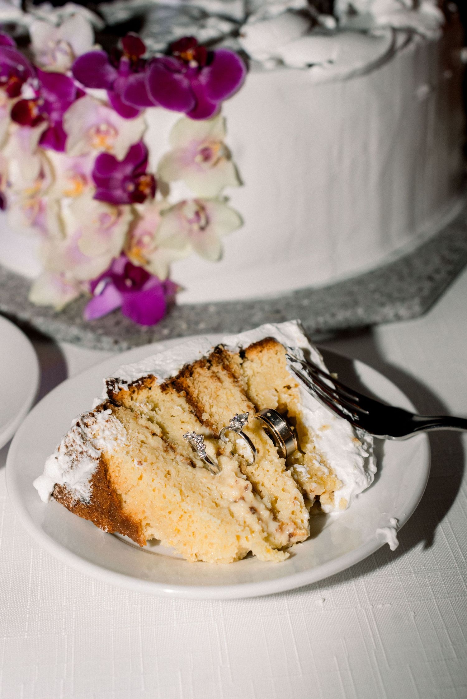 A slice of wedding cake with purple and white orchid flowers decorating the top tier on a silver platter.