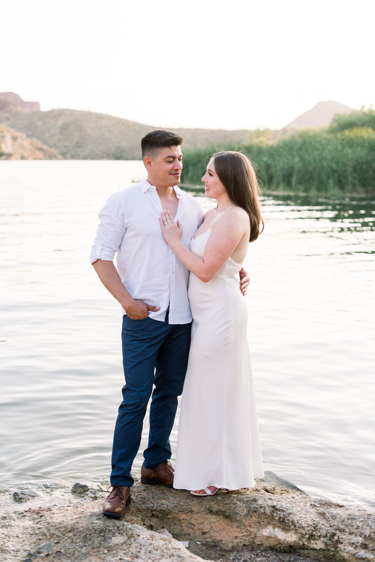 A romantic couple poses for engagement photos on rocks by a serene lake during golden hour.