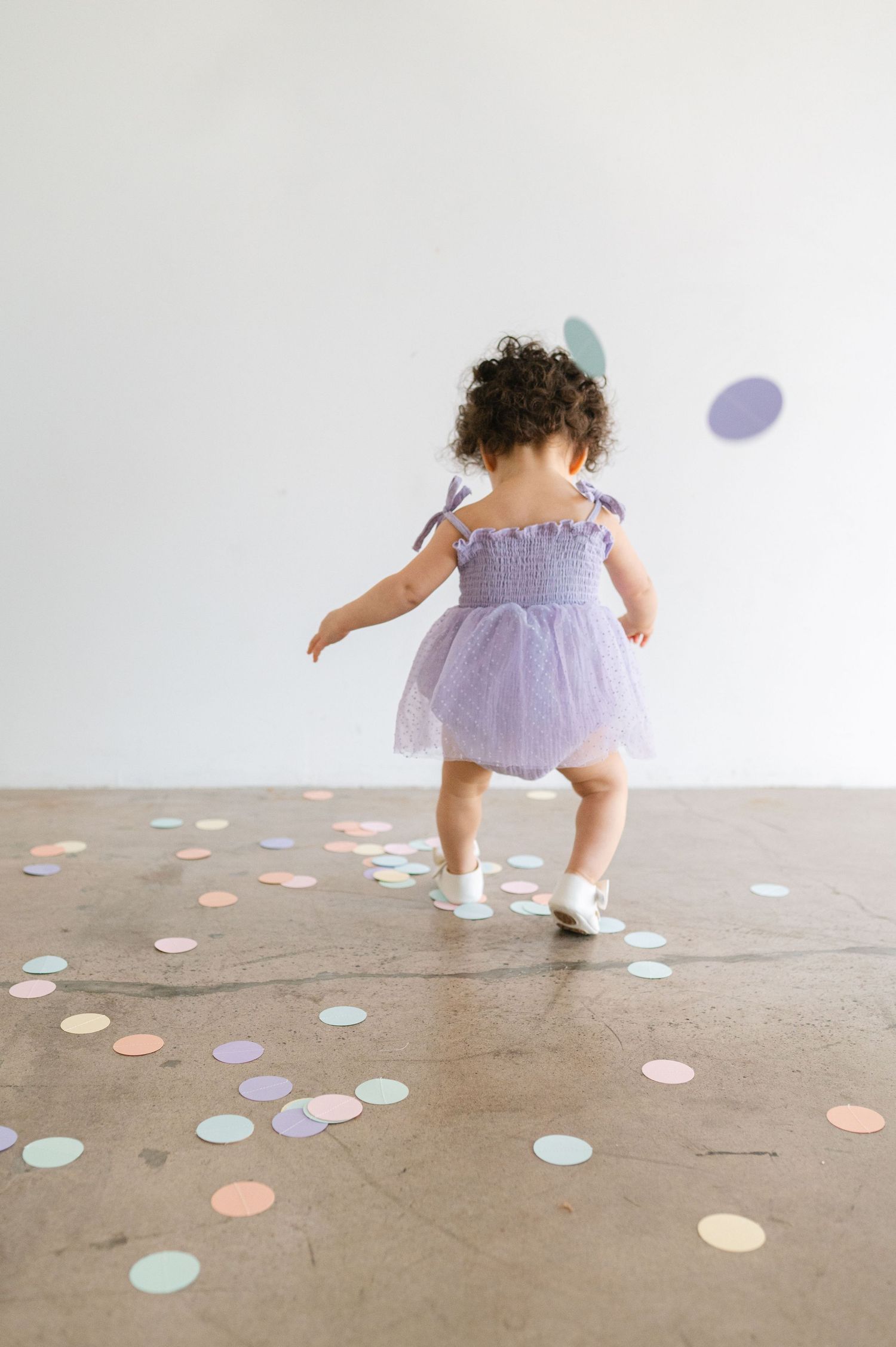 Small child in purple dress dances and twirls among colorful paper dots scattered across a concrete floor.