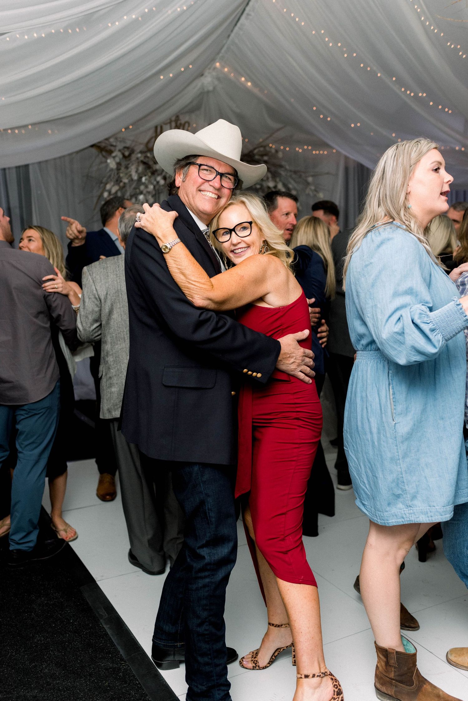 Party guests dancing and embracing on white dance floor under draped fabric ceiling with twinkle lights.