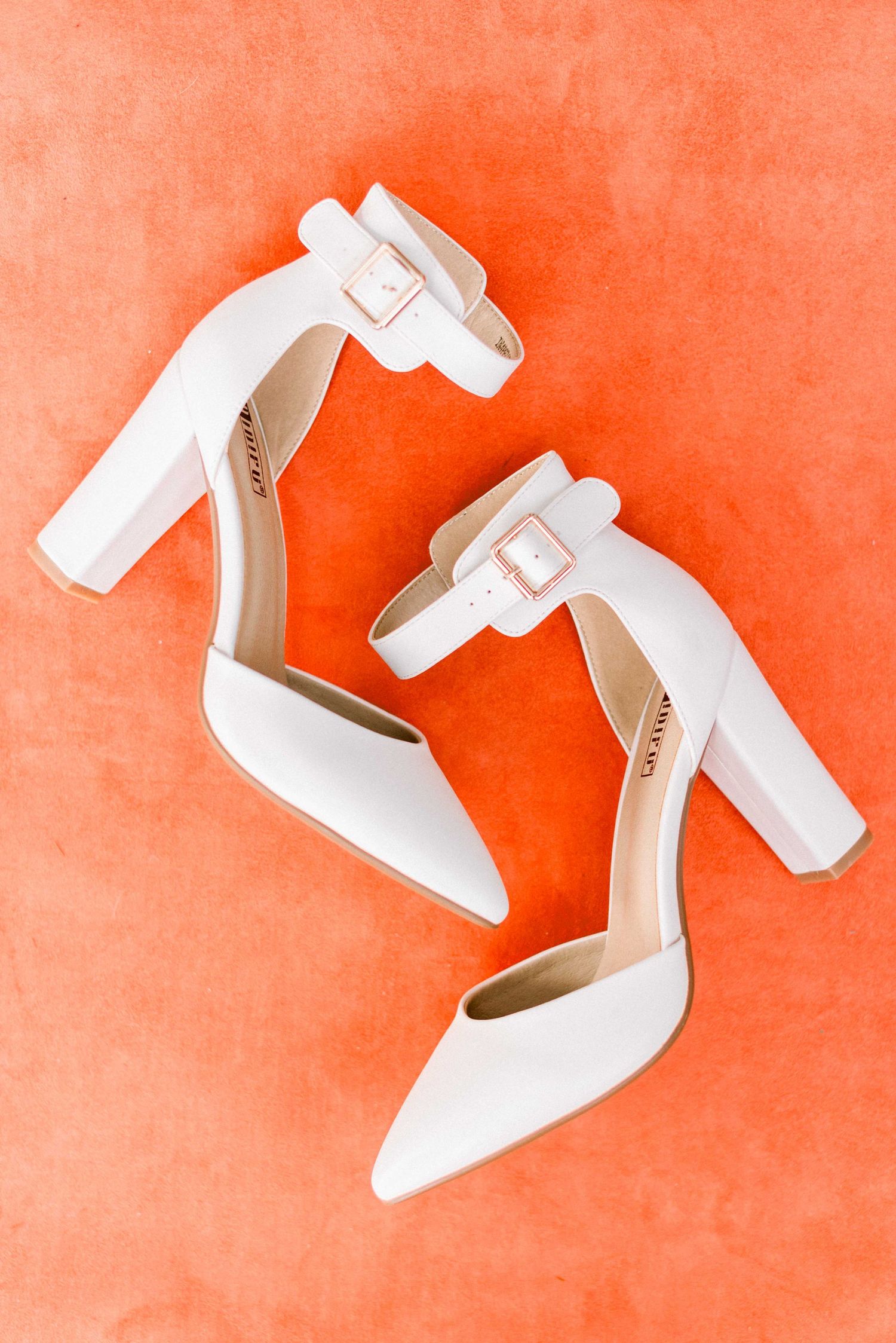 Elegant white wedding heels with ankle straps photographed on coral background.