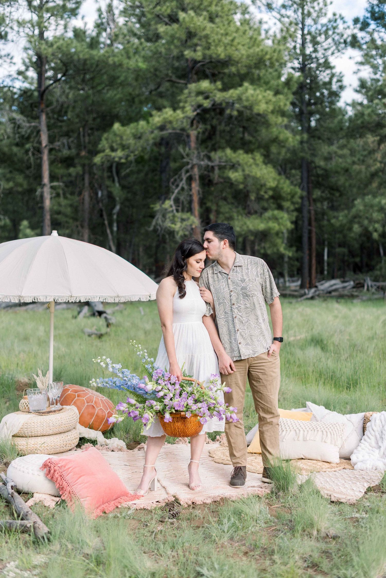 Romantic outdoor scene with couple standing by umbrella and floral arrangements in pine forest.